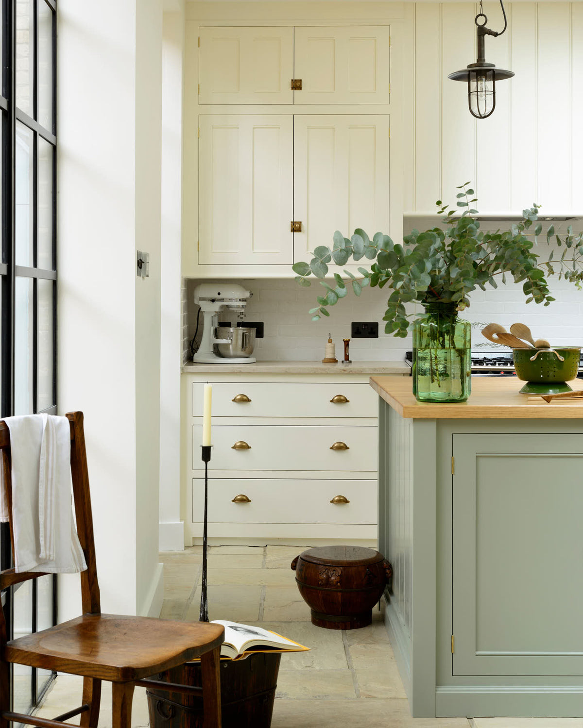 A quite corner of a bright and spacious classic English kitchen features a long white cooker run with wall cupboards and a pale-blue kitchen island with a solid oak worktop. Adorned with metal embellishments like industrial pendant lights, a silver mixer, a jade-green vase filled with a floral arrangement, and vintage wooden furniture, it creates a wonderful mix of simplicity and charismatic personality.