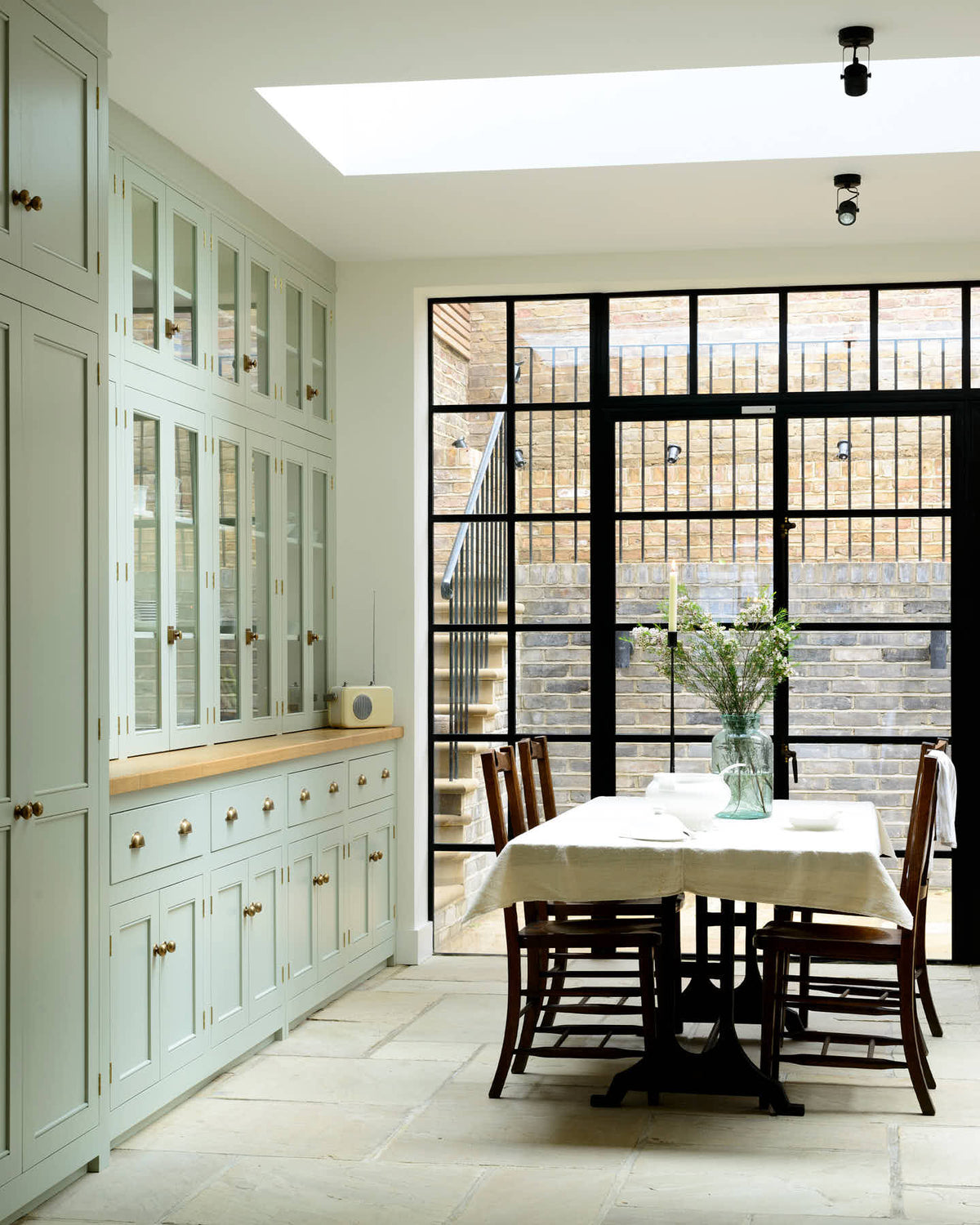 This bright and spacious classic English kitchen features a full-height cupboard, a long pantry run with glazed counter cupboards, and a solid oak worktop. Adorned with a vintage cream radio, an aqua-green vase filled with a floral arrangement, and vintage wooden furniture, it creates a delightful mix of simplicity and charismatic personality.