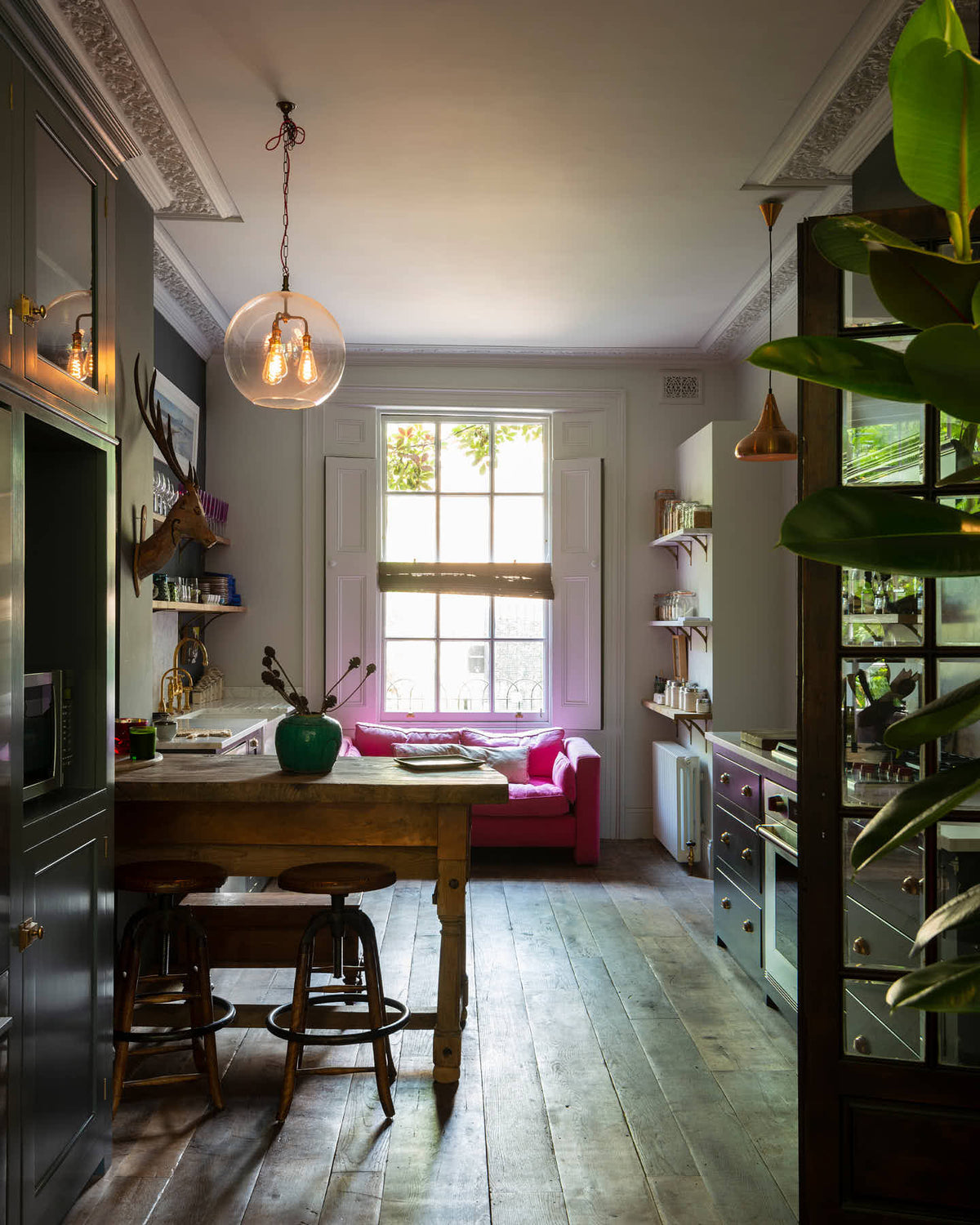 An eclectic, navy blue, classic English kitchen features a short and humble sink run, cooker run and a freestanding cabinet. The long, corridor-like space of a Georgian townhouse is beautifully decorated with beloved vintage items and furniture, paintings, copper pendant lights and unique kitchen equipment, giving the room a lot of whimsy, character and functionality.