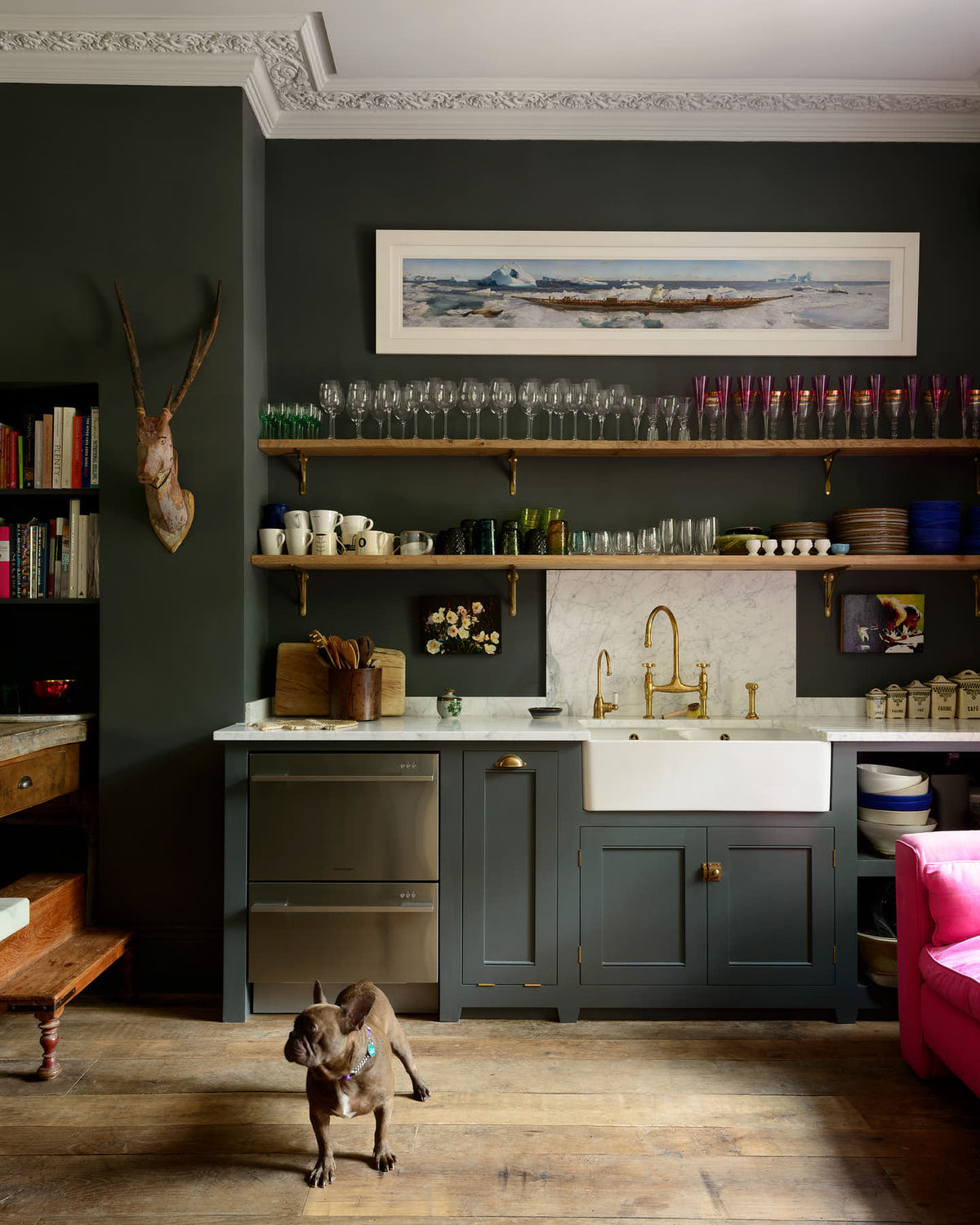 An eclectic, navy blue, classic English kitchen features a short and humble sink run with a double Belfast sink, an aged brass Ionian tap, a Carrara marble worktop, and a square splashback at the centre. The long, corridor-like space is beautifully decorated with beloved vintage items and furniture, paintings, unique kitchen equipment and a cute small dog, giving the room a lot of whimsy, character and functionality.