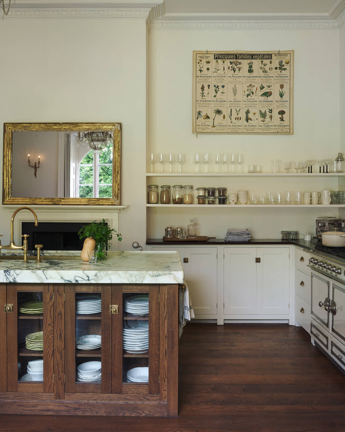 A grand yet simple white linen classic English kitchen, featuring white and cream splashback tiles, a cooker run with a polished sapele worktop, and a kitchen island topped with Arabescato marble, a polished brass sink, and an aged brass Ionian tap. The space is decorated with a large scientific-style illustration of different vegetable species and a large metal-and-crystal chandelier.