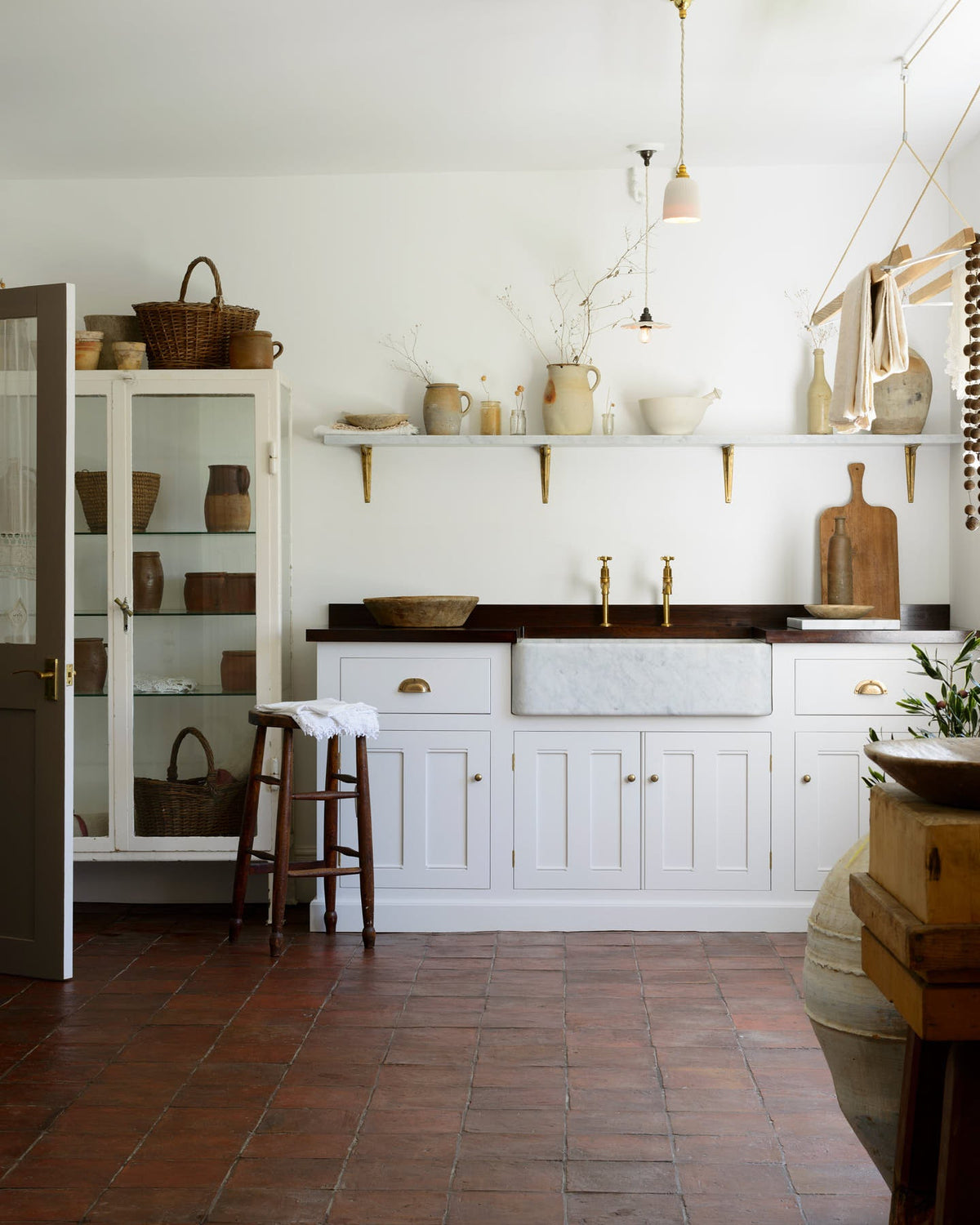 The simple and functional linen white scullery room with a modest sink run with polished sapele, a large single Carrara marble sink, and aged brass Mayan taps. The warm reds and browns of the terracotta square tiles, vintage furniture, ceramics, woven baskets and a deVOL laundry maid serve the room with both a purpose and beautiful aesthetics.