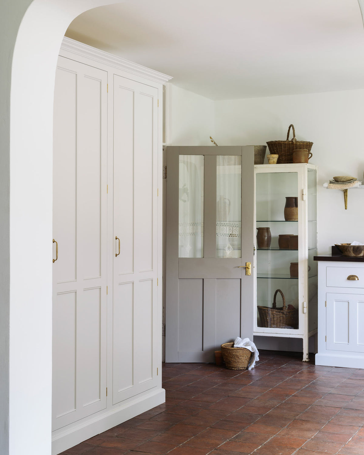An angled view of a simple and functional linen white scullery room with a modest sink run with polished sapele, a large single Carrara marble sink, and aged brass Mayan taps. The warm reds and browns of the terracotta square tiles, vintage furniture, and ceramics serve the room with both a purpose and beautiful aesthetics.