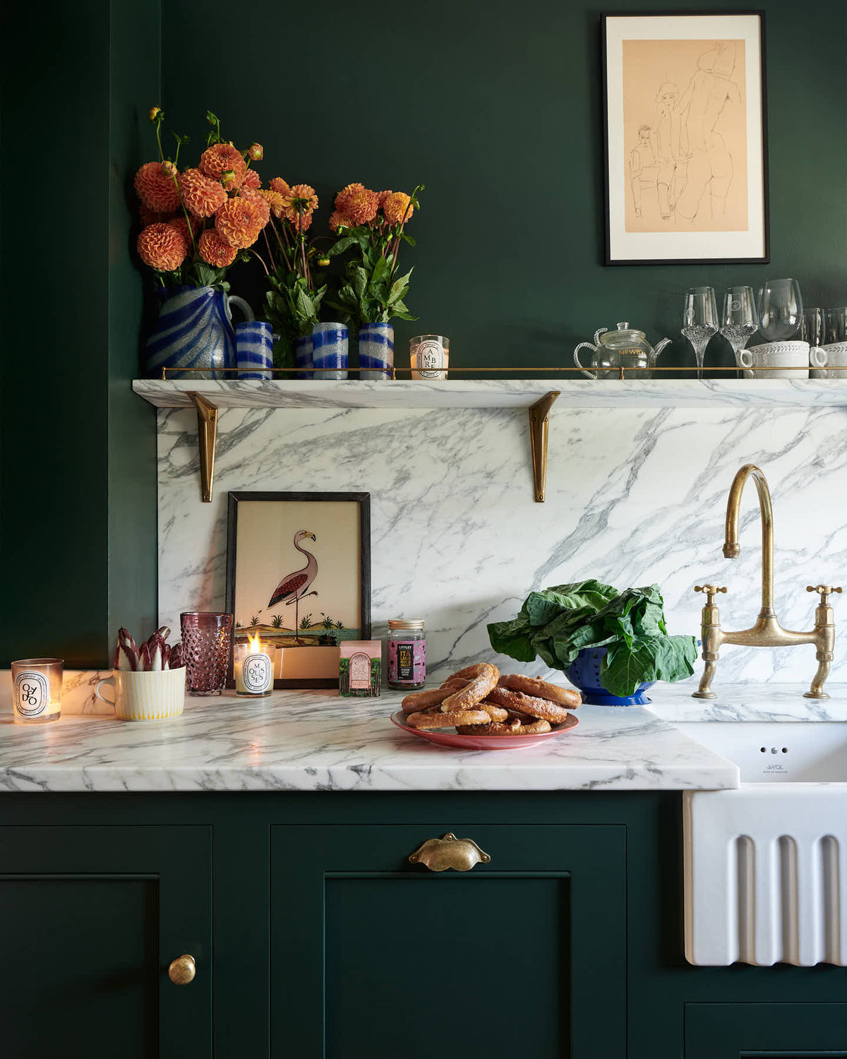 A close-up of a small but luxurious Bakehouse green classic English kitchen featuring a sink run with an Arabescato worktop, splashback and counter shelf, a single fluted Belfast sink and an aged brass Ionian tap. Beautifully decorated with all sorts of colourful and intricate bric-a-brac, framed illustrations, a plate of pretzels, washed lettuce in a blue strainer, and red chicory in a large mug, giving the kitchen both functionality and personality.