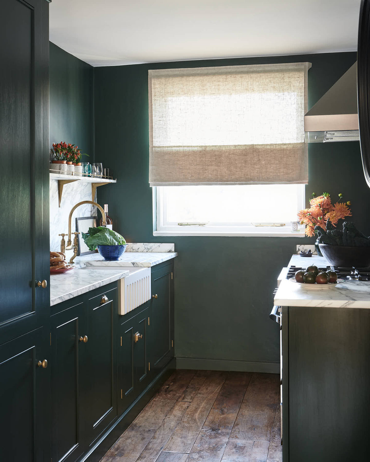 An angled view of a small but luxurious Bakehouse green classic English kitchen featuring a sink run with an Arabescato worktop, splashback and counter shelf, a single fluted Belfast sink and an aged brass Ionian tap. Beautifully decorated with all sorts of colourful and intricate bric-a-brac, framed illustrations, a plate of pretzels, washed lettuce in a blue strainer, and red chicory in a large mug, giving the kitchen functionality and personality.