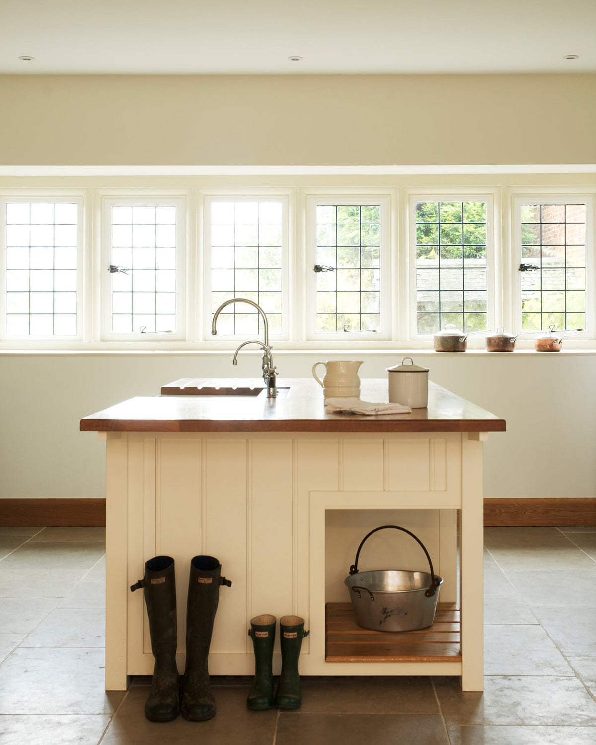 This simple and spacious classic English kitchen features a kitchen island equipped with a double Belfast sink and an antique silver Ionian tap atop a solid oak worktop. Adorned with a vintage metal basket, simple ceramics, copper pans, and boots, it creates a humble and functional family kitchen.