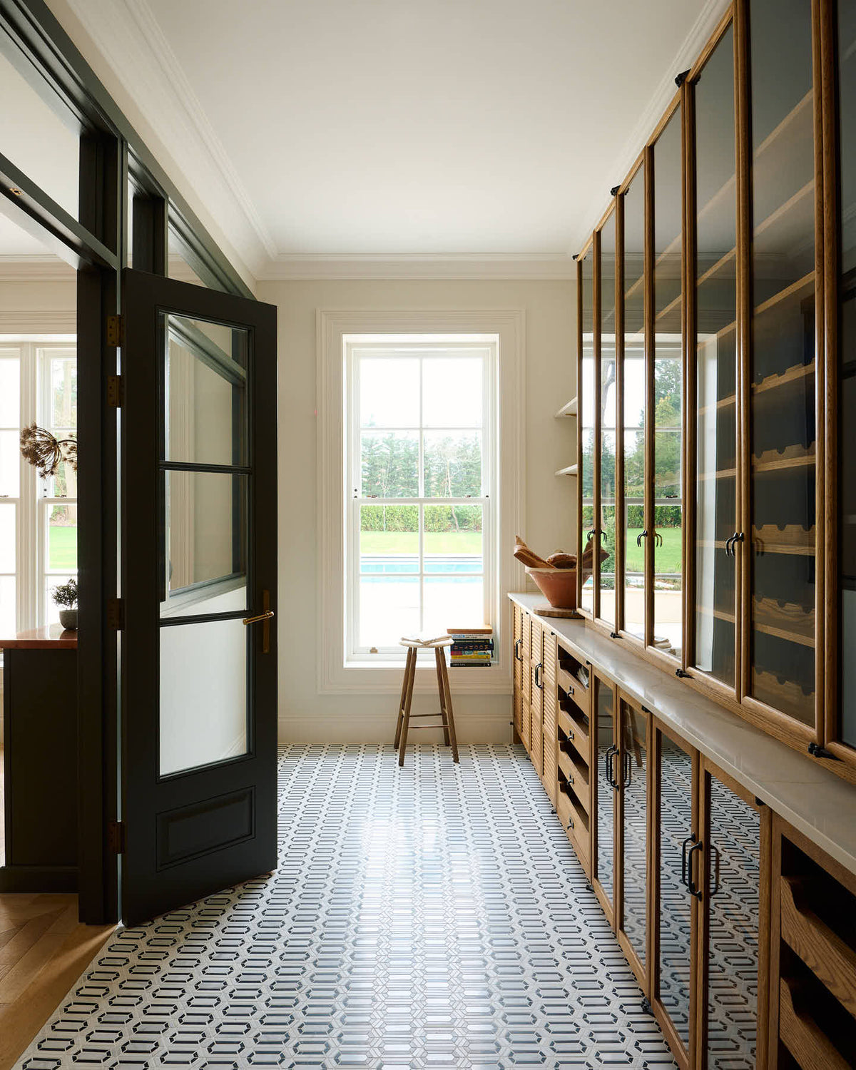 One side of a grand, earth-toned classic kitchen features a Silestone worktop cooker run, glazed wall cupboards, a full-height pantry, a copper canopy, a large island with a copper worktop, and a ladder. Adorned with warm colours such as an aged brass hanging rail, deVOL Heirloom gaselier lights, textured woven baskets, vintage ceramics and loaves of bread, it makes a spectacular use of space but also down-to-earth.