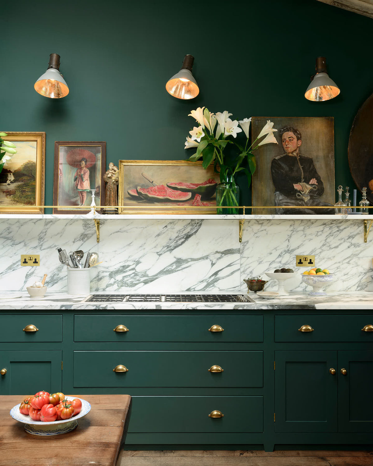 A lush green, classic English kitchen featuring an L-shaped sink and cooker, all complemented by Arabescato marble for the worktop, splash back, and countertop shelf. An angled wooden skylight window allows natural light to flow into the room, highlighting white lilies in glass vases, a rich oak dining table, framed antique paintings, and industrial-style gas light fixtures on the walls.