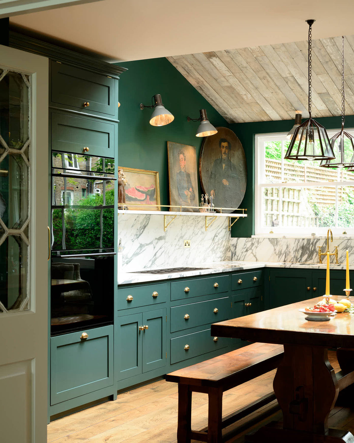 A lush green, classic English kitchen featuring an L-shaped sink and cooker, all complemented by Arabescato marble for the worktop, splashback, and countertop shelf. The kitchen includes full-height cabinets and a wall cupboard, creating a luxurious yet bohemian atmosphere. An angled wooden skylight window allows natural light to flow into the room, highlighting a rich oak dining table with a matching bench, framed antique paintings, and industrial-style gas light fixtures on the walls.