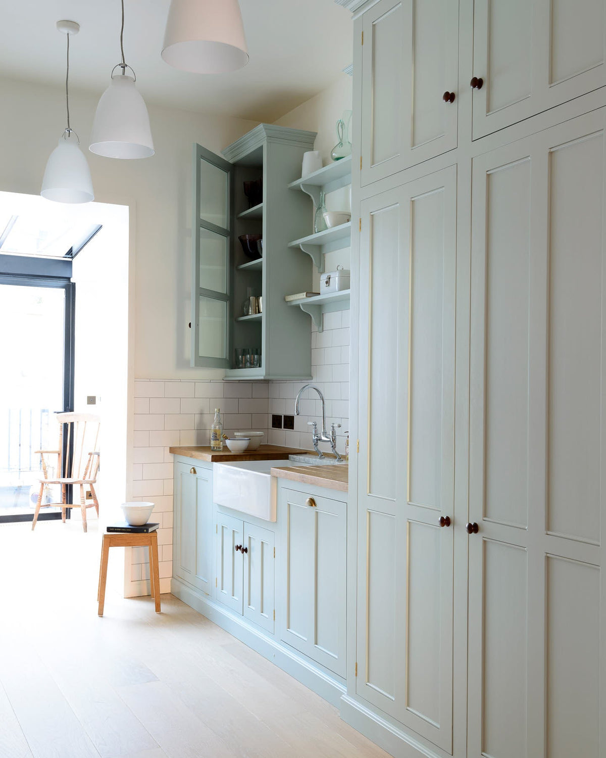 A close-up of a simple and stylish classic English kitchen’s double Belfast sink with antique silver Ionian tap, Carrara marble worktop, glazed wall cupboard, and shelf. Beautifully decorated with light copper rimmed clock, glass containers, wooden cleaning equipments, and white splash back tiles, it creates a functional and beautiful kitchen space.