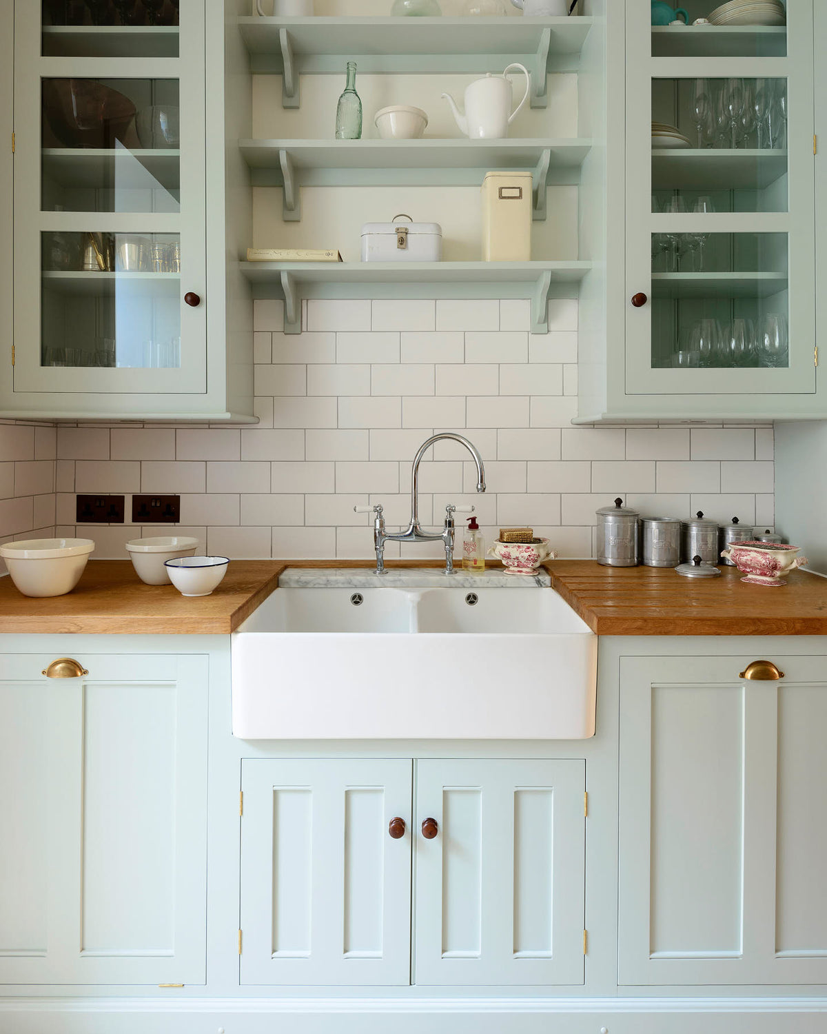 A close-up of a pale-blue classic English galley kitchen features a sink run with a solid oak worktop, a double Belfast sink, a chrome Ionian tap, glazed wall cupboards, pantry cupboards, and shelves. Adorned with artisan ceramics, vintage containers, and simple platters, it creates a humble and practical kitchen that embraces vintage 80s aesthetics.