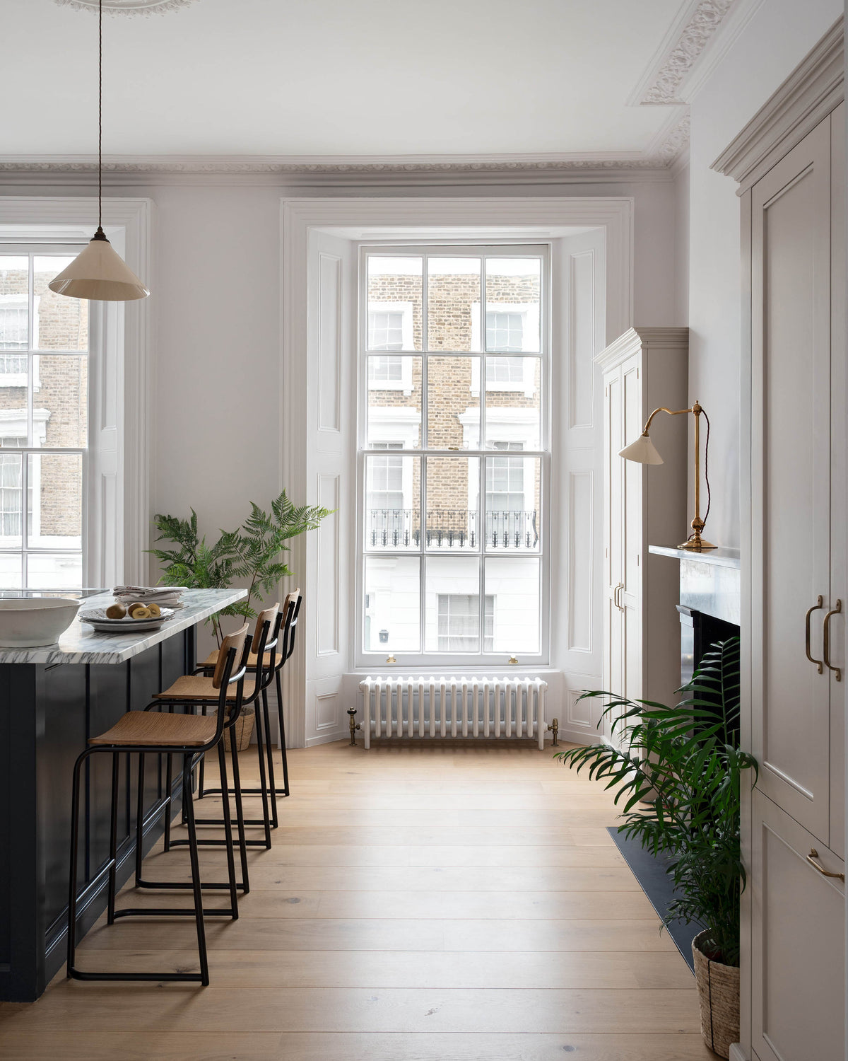 A humble yet elegant off-white Classic English kitchen features a Pantry blue kitchen island with a cooker, full height cabinets, and plumber’s stools. Adorned with artisan ceramics, a plate of fresh kiwis, a creamware pendant light, a grand tour desk lamp, and house plants, creating an intimate social and practical kitchen space.