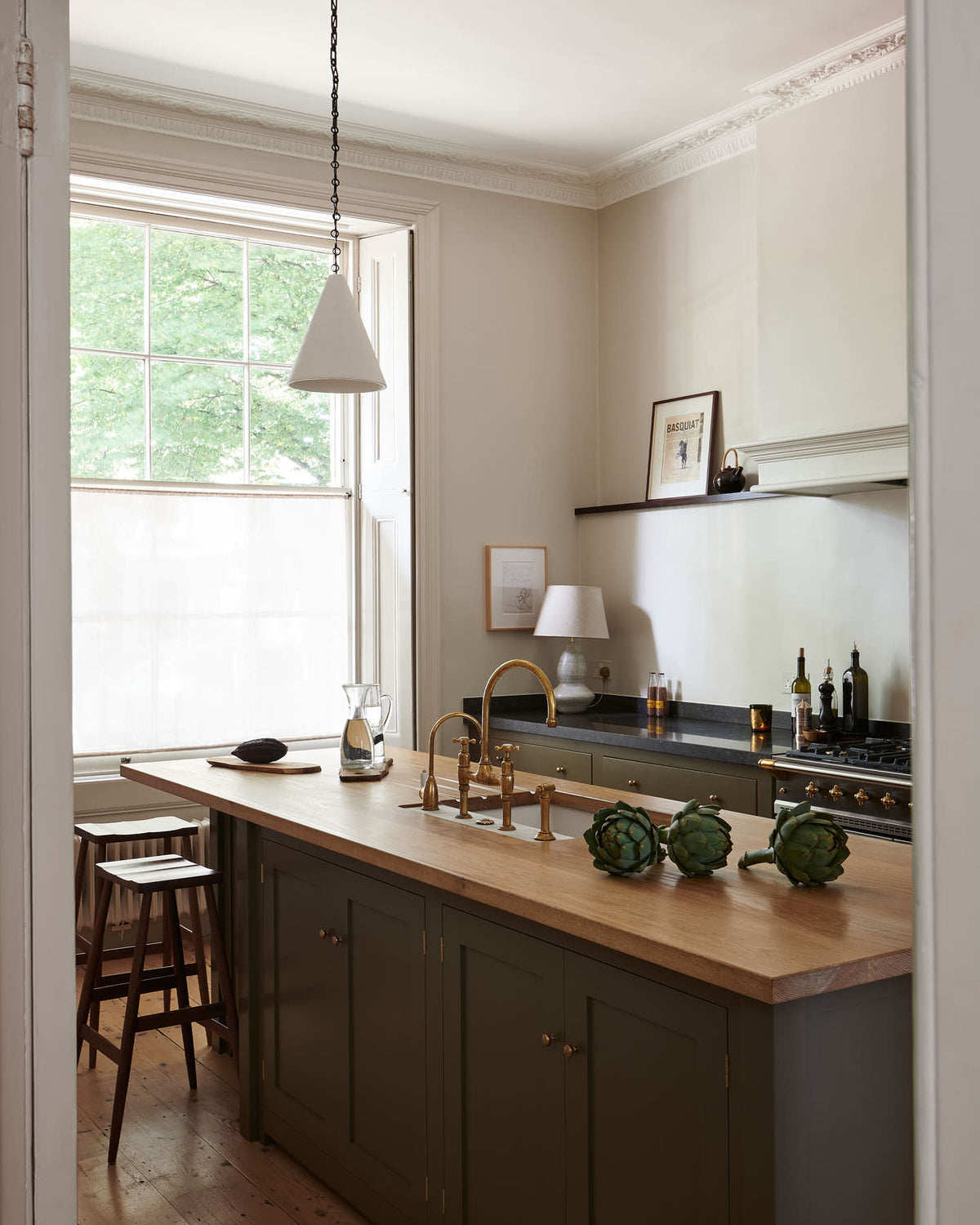 Looking through a white open door, one sees an elegant olive-brown classic kitchen featuring a cooker run topped with Belgian blue limestone, a Shaker canopy, and a built-in shelf. At the centre is a kitchen island with a sink, a solid oak worktop, an aged brass Ionian tap, and a breakfast nook with wooden stools at the far edge. The tall-ceilinged room is decorated with slender bottles of wine and vinegar, a pepper grinder, a vibrant green artichoke, and small, delicate framed illustrations.