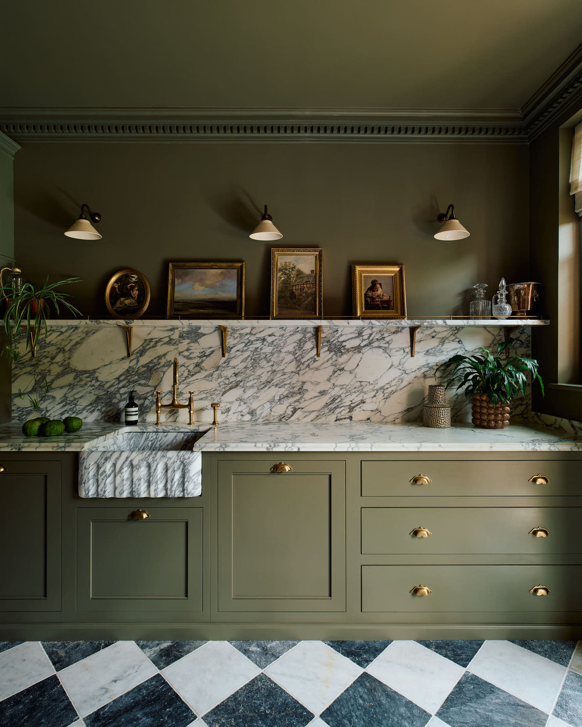 A grand and atmospheric olive green classic English kitchen features a marble worktop, splashback, and shelf along the sink run, a single fluted marble sink and an aged brass Ionian tap. Three beautiful Heirloom wall lights hang above gold-framed famous paintings, potted plants, and eccentric liquor bottles.