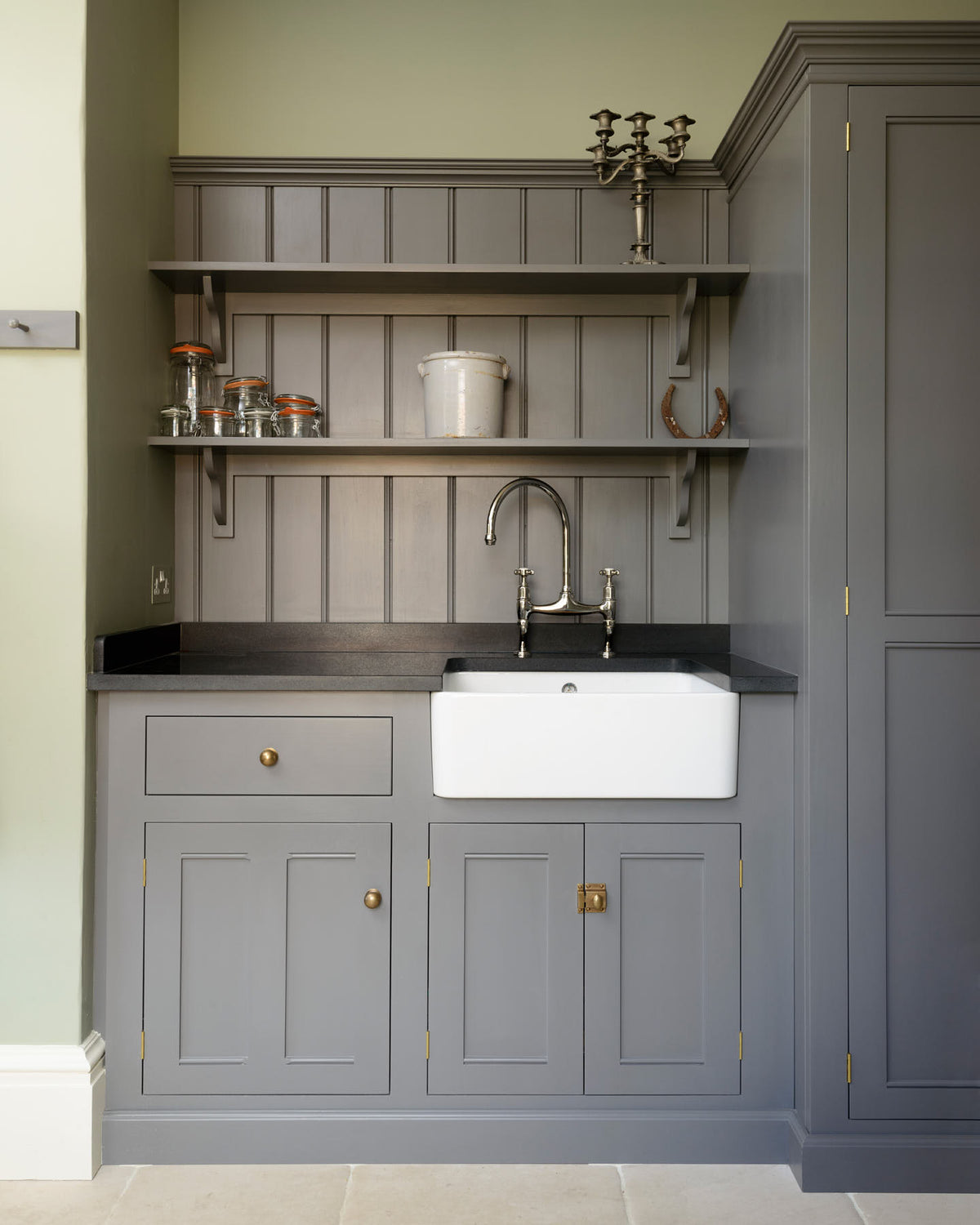 A closer look at a muted-grey boot room showcases a small sink run featuring a single Belfast Ionian sink, an antique silver Ionian tap, and a black granite worktop, complemented by shelves and full-height storage cupboards. Beautifully decorated with a vintage candelabra, a horseshoe, a beloved ceramic piece, and empty glass jars, it creates a practical and family-oriented space.