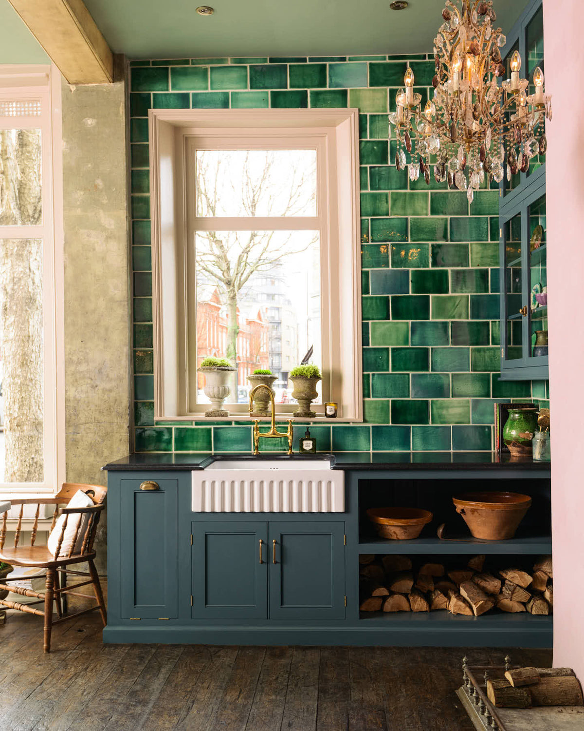 The playful pastel yet classic kitchen features a Clerkenwell blue sink run with a black granite worktop, emerald green splash back tiles, a single fluted Belfast sink, and an aged brass Ionian tap. A long kitchen island with a breakfast nook and deVOL bum stools adds charm. Decorated with chopped firewood, vintage ceramics, and a crystal chandelier, this kitchen has both functionality and personality.