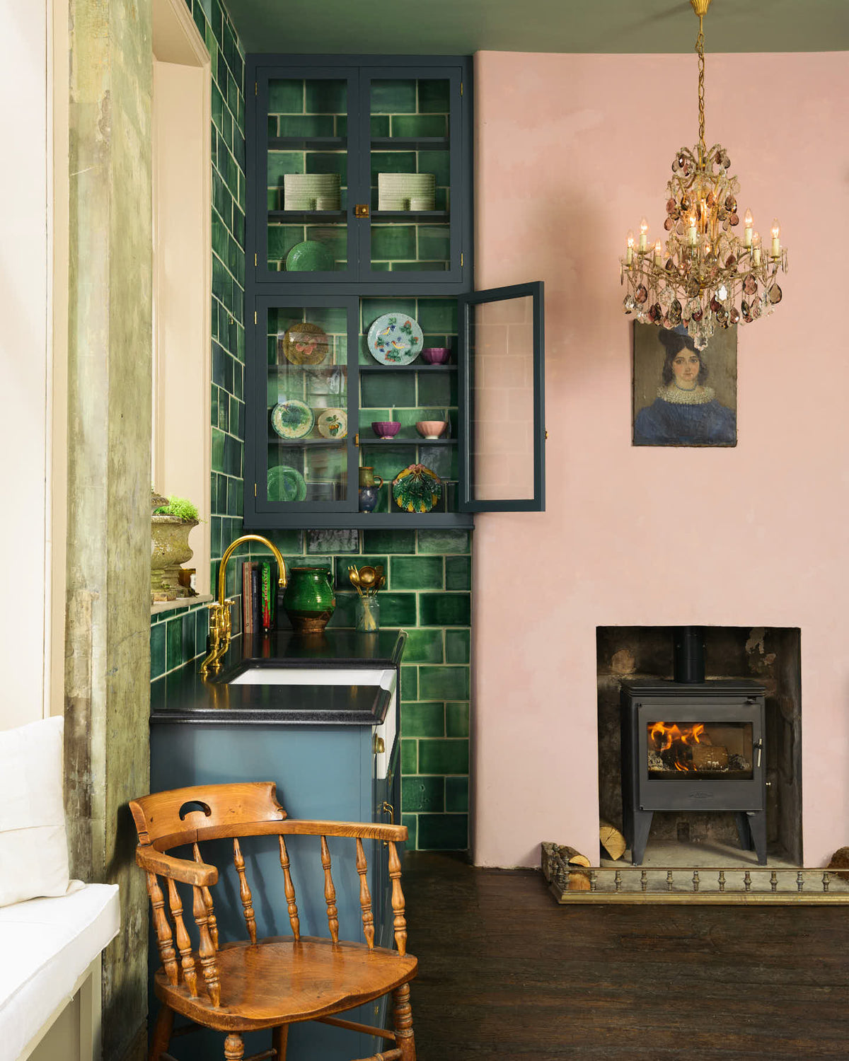 The playful pastel yet classic kitchen features a large Clerkenwell blue full-height pantry cupboard with a black granite worktop, emerald green splashback tiles, and a pink chimney with a small iron wood stove. Decorated with chopped firewood, vintage ceramics, a small oil painting portrait and a crystal chandelier, this kitchen has both functionality and personality.