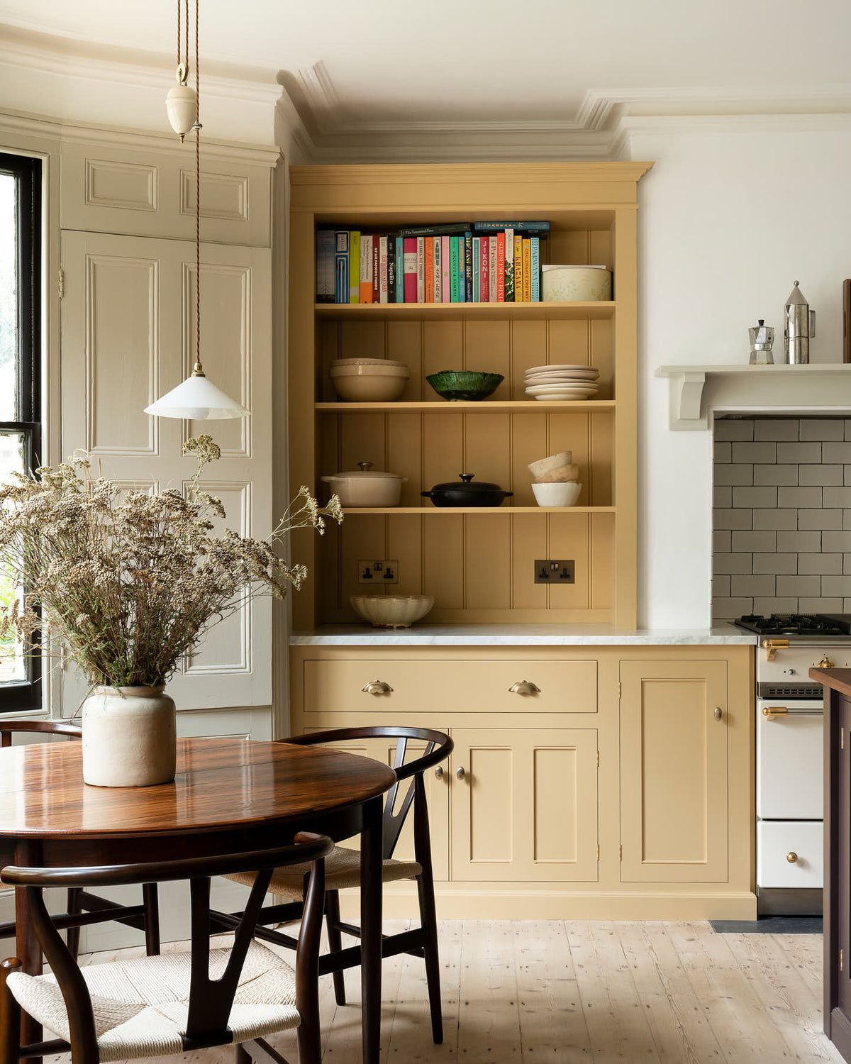 A lovely and simple blush yellow, classic English kitchen features a cooker run with Carrara marble worktop, two open countertop cupboards, a white gas stove inside a chimney breast, porcelain splashback, a purple-brown kitchen island with a solid oak worktop and aged brass Mayan taps. A circular coffee table with matching chairs, a deVOL gaselier light, and a collection of muted ceramics and tall wildflowers create an elegant and inviting atmosphere in the room.