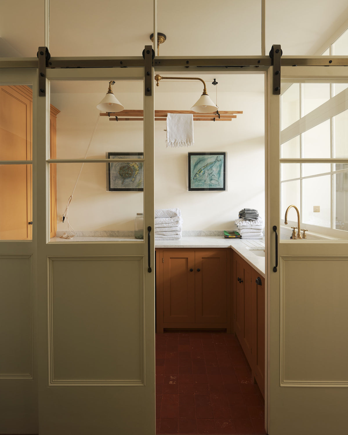 A cream sliding door leads to a scullery yellow laundry room featuring an L-shaped sink with a white marble countertop and a stunning aged brass Ionian faucet. Neatly stacked white towels are arranged in layers, and two framed artworks adorn the walls. In the centre of the ceiling hangs a deVOL Gaselier light, while a deVOL Laundry Maid, with a towel draped over it, is positioned behind.