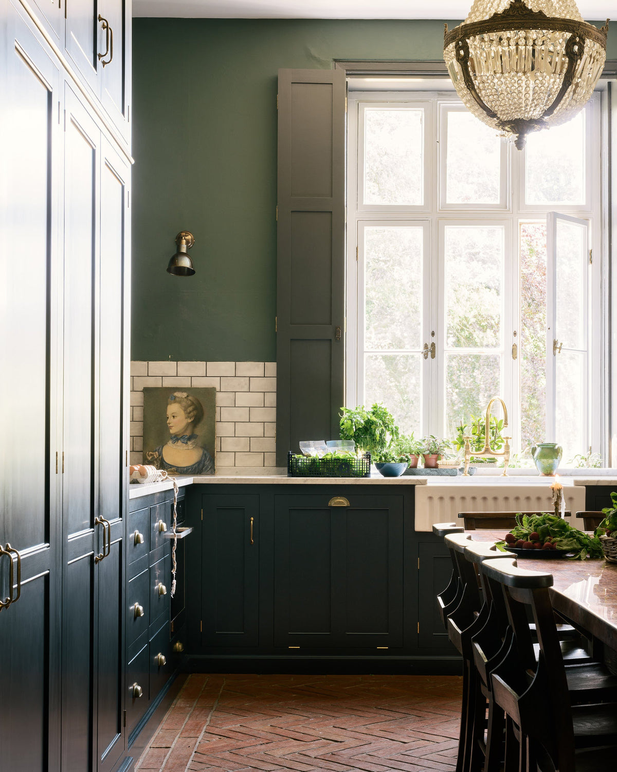 A grand L-shaped kitchen with bespoke Georgian-style cabinetry, painted in a dark Prussian blue and decorated with traditional brass fittings. The room has a very high ceiling and tall painted shutters on either side of a beautiful original window. The walls are a soft moss green with a band of mottled white metro tiles. A large antique crystal chandelier hangs above a rustic herringbone brick floor.
