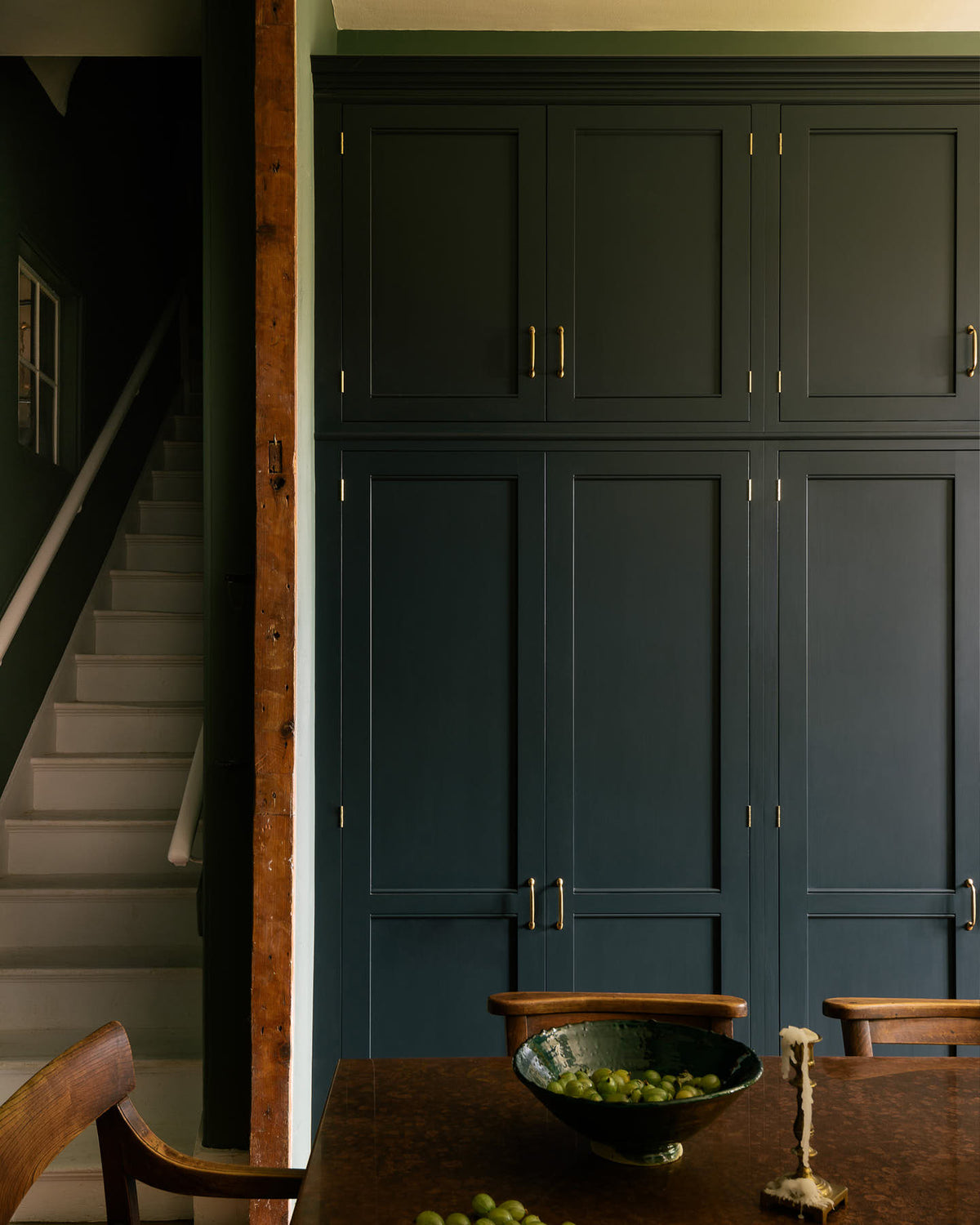 A grand L-shaped kitchen with bespoke Georgian-style cabinetry, painted in a pantry blue and decorated with traditional brass fittings. The room has a high ceiling and tall painted shutters on either side of a beautiful original window. The walls are a soft moss green with a band of mottled white metro tiles. A large antique crystal chandelier hangs above a rustic herringbone brick floor.