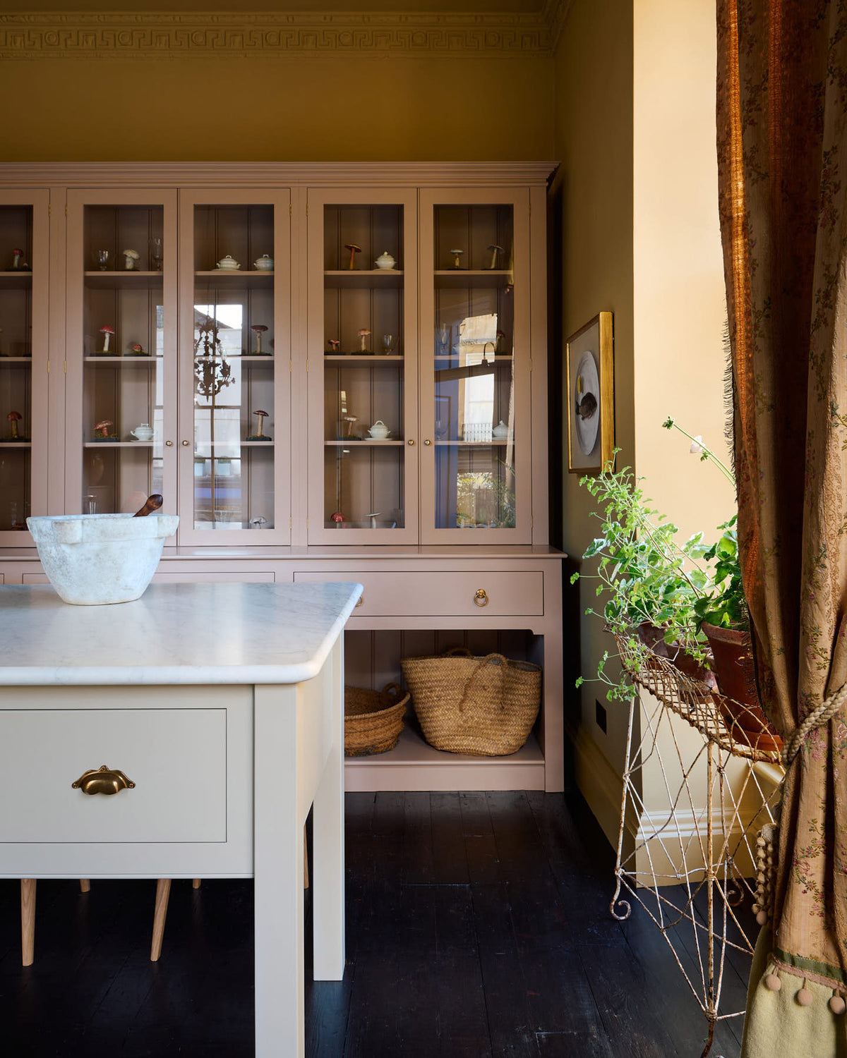 A corner of a warm yellow room with a large Princelet pink dresser showcasing glazed cupboards filled with delicate ceramics, drinking glasses, and papier-mâché mushrooms, giving the space a relaxed, living-room feel. The space features a spacious prep table, large ceramic bowls, and a golden-framed painting depicting a fish and a lemon.