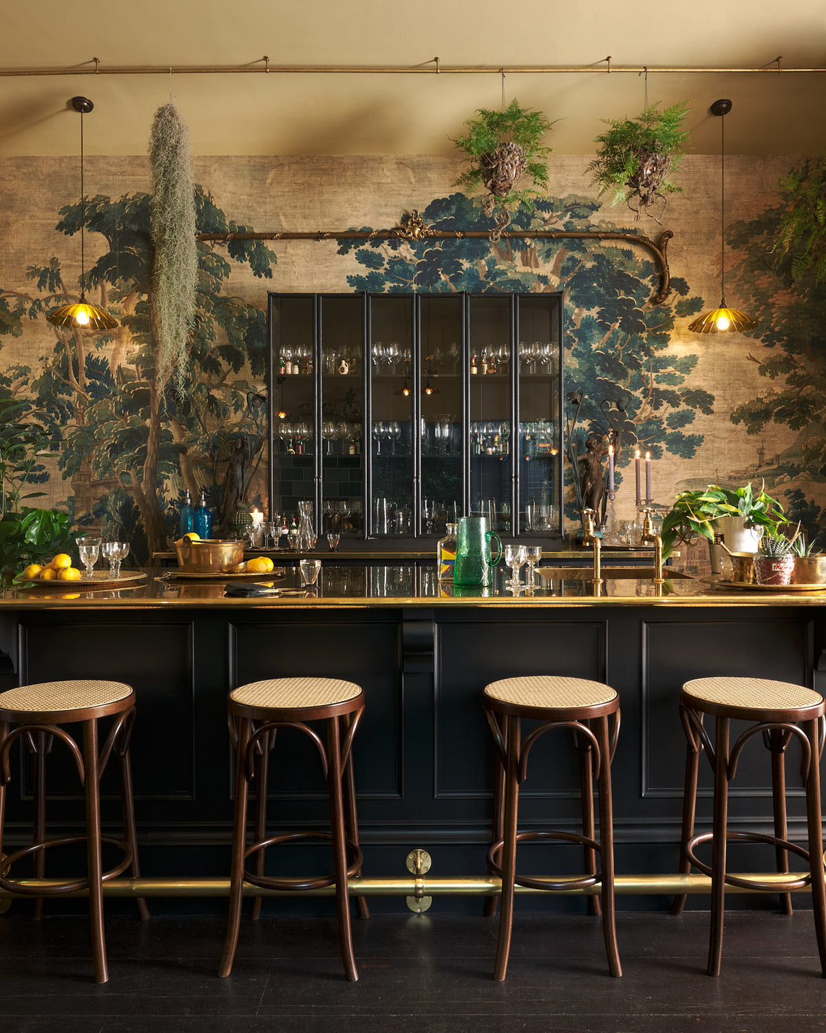 A chic and glamorous Bobbin Black Haberdasher’s bar featuring a kitchen island with a polished brass worktop, a single polished brass sink, aged brass Mayan taps, a brass foot rail, glazed base cabinet with internal drawers, tall countertop cupboards, and woven top bentwood café stools. Set against an illustrated foliage wallpaper and adorned with hanging plants, wine glasses, and vintage decanters, it creates a beautiful and intimate personal bar.