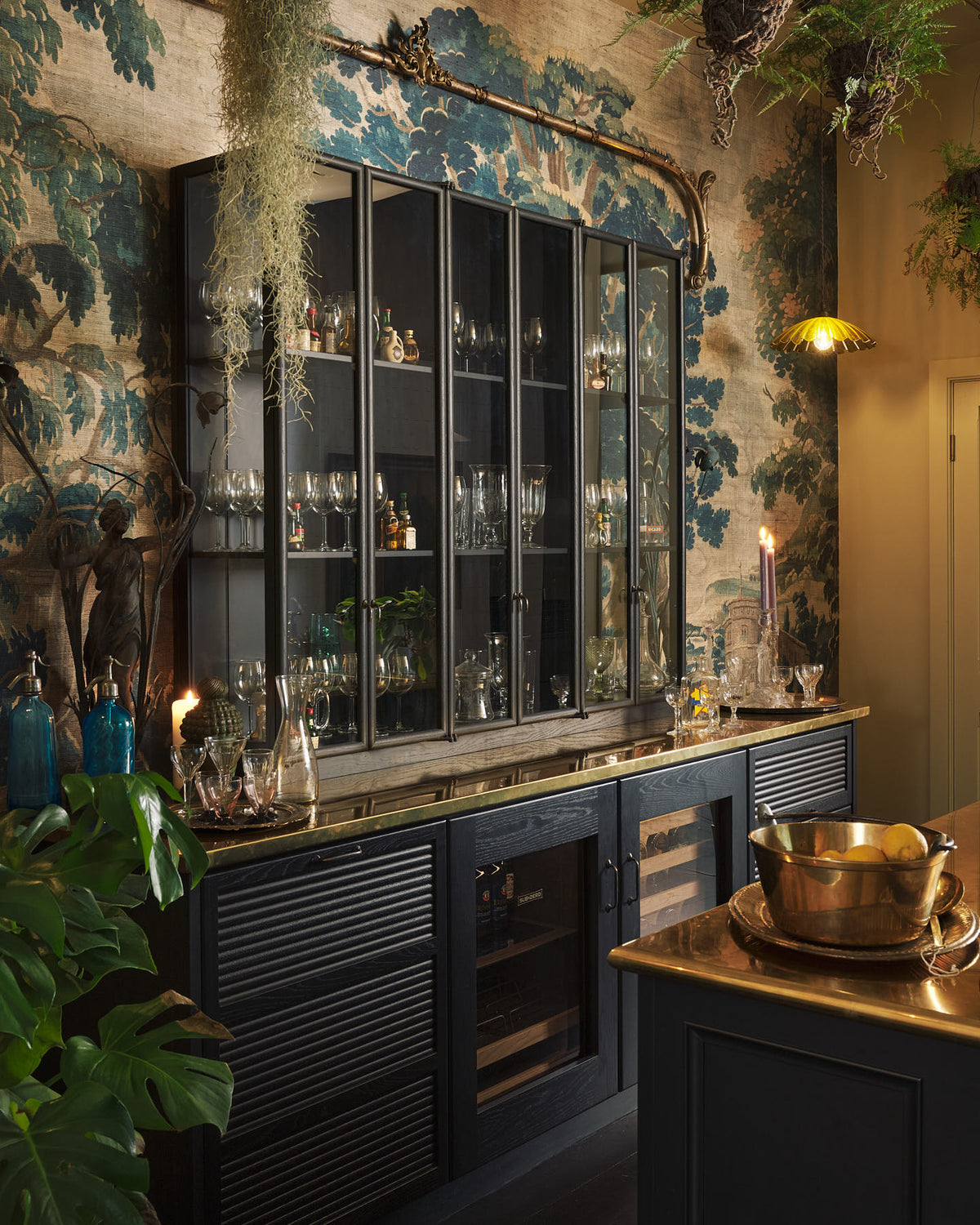 An oblique view of a chic and glamorous Bobbin Black Haberdasher’s bar featuring a kitchen island with a polished brass worktop, glazed base cabinet with internal drawers, and tall countertop cupboards. Set against an illustrated foliage wallpaper and adorned with hanging plants, wine glasses, and vintage decanters, it creates a beautiful and intimate personal bar.