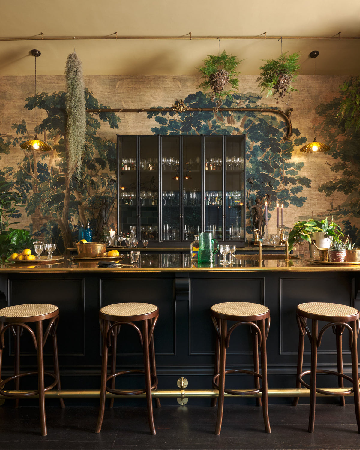 A chic and glamorous Bobbin Black Haberdasher’s bar featuring a kitchen island with a polished brass worktop, a single polished brass sink, aged brass Mayan taps, a brass foot rail, glazed base cabinet with internal drawers, tall countertop cupboards, and woven top bentwood café stools. Set against an illustrated foliage wallpaper and adorned with hanging plants, wine glasses, and vintage decanters, it creates a beautiful and intimate personal bar.
