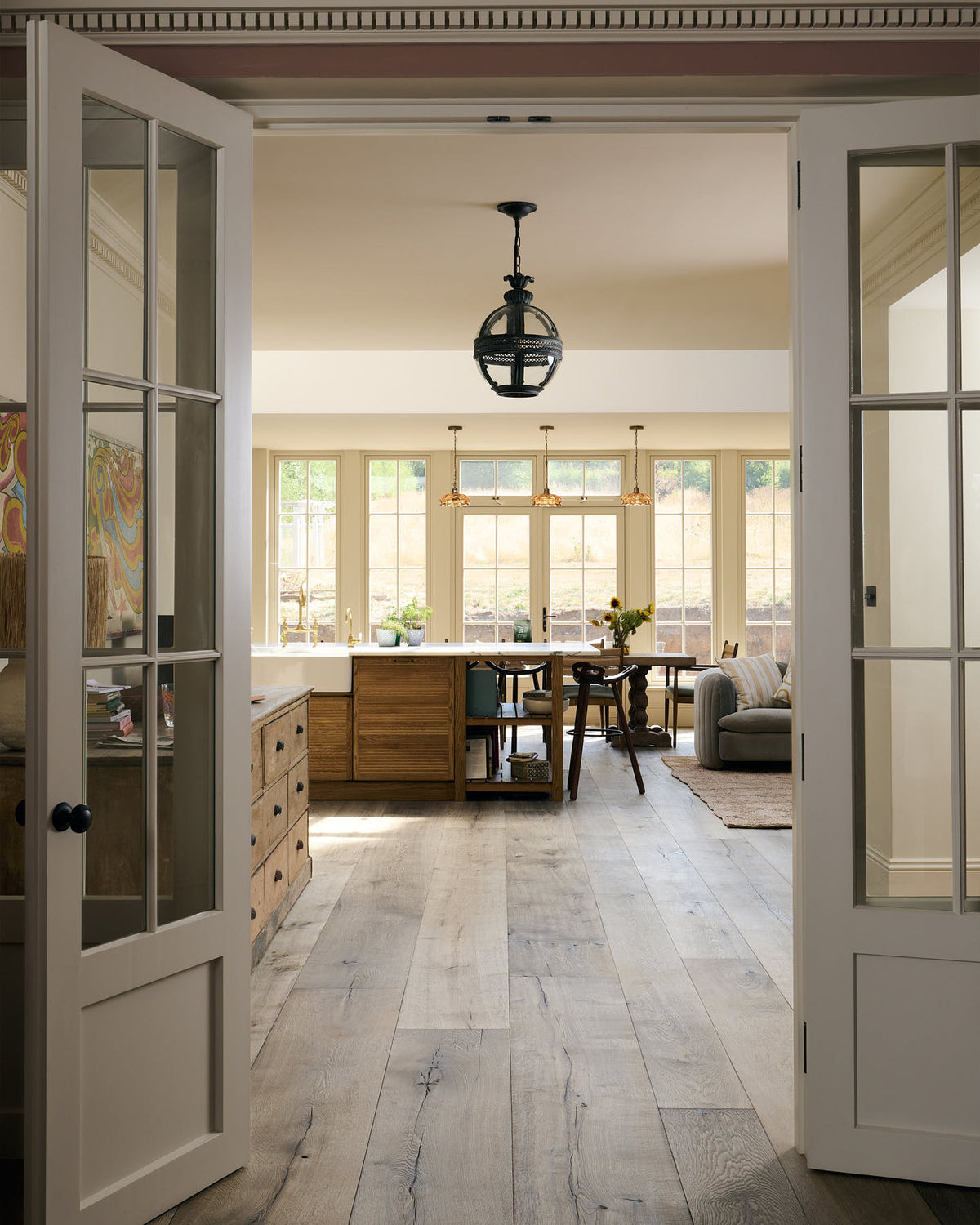Past white French doors is an elegant and spacious Haberdasher’s kitchen features a large cooker run, a kitchen island with a double Belfast sink, an aged brass Ionian tap, an Arabescato marble worktop, splashback, and glazed cupboards. Adorned with vintage furniture, artisan ceramics, glass flower pendant lights, and a sunflower arrangement, this kitchen creates a classic social space with functional storage.