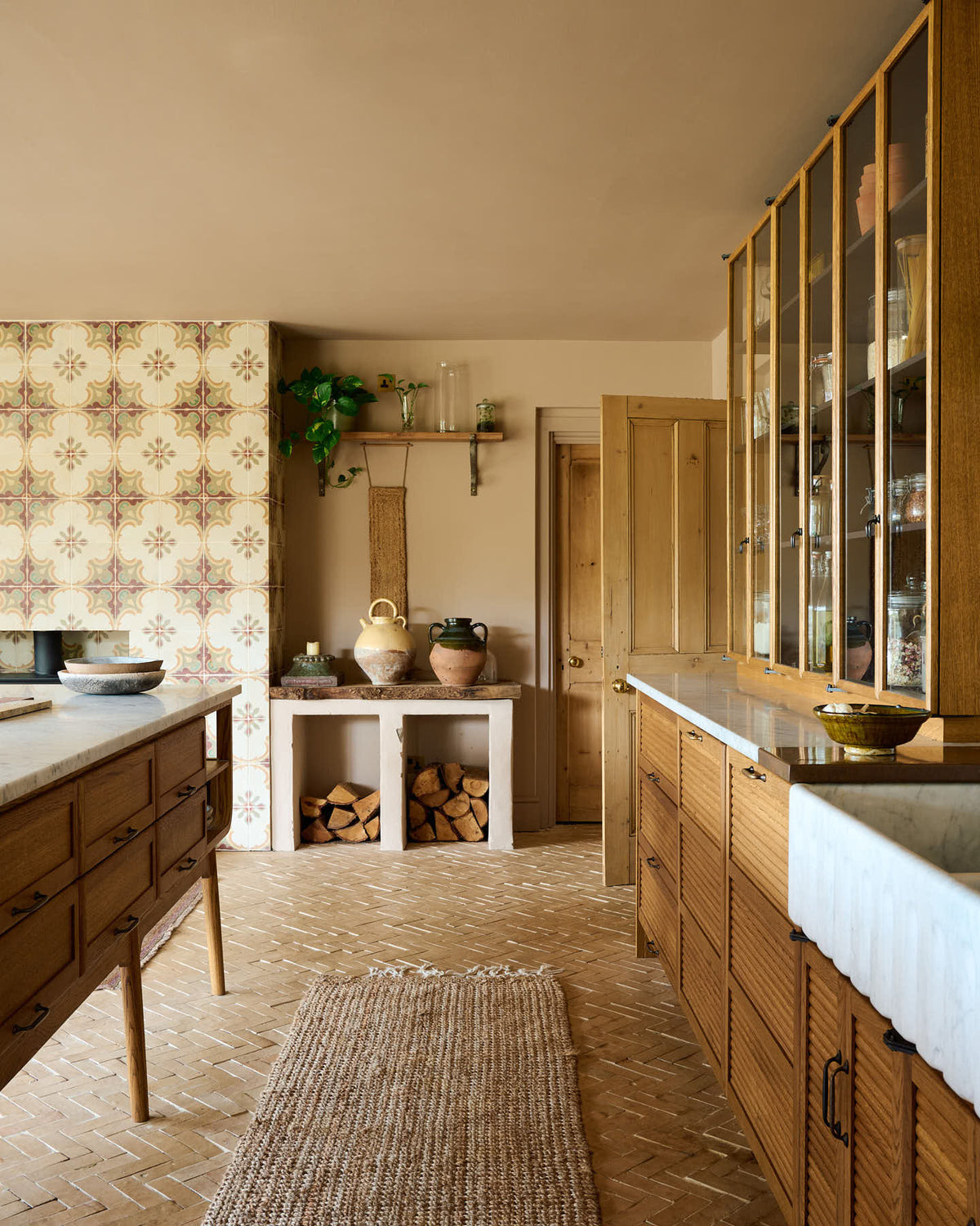Nestled between a warm and chic Haberdasher’s kitchen features an L-shaped sink and cooker run, countertop cupboards, a kitchen island, a Carrara and copper mix worktop, and a double-fluted marble sink, is a firewood storage area. Adorned with artisan ceramics, a large vintage rug, and luscious green house plants, this kitchen creates a warm and inviting Spanish-inspired space in Yorkshire.