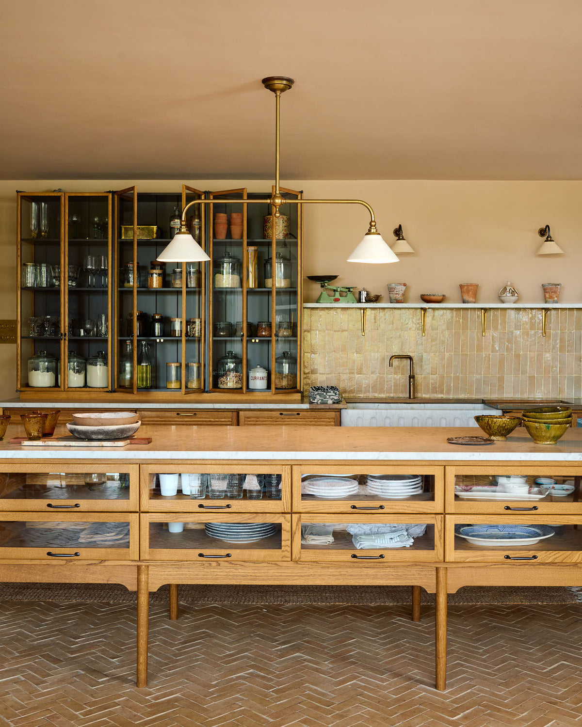 A warm and chic Haberdasher’s kitchen features an L-shaped sink and cooker run, countertop cupboards, a kitchen island, a Carrara and copper mix worktop, and a double-fluted marble sink. Adorned with artisan ceramics, a deVOL gaselier light, Heirloom wall lights, large vintage rugs, and engraved drinking glasses, this kitchen creates a warm and inviting Spanish-inspired space in Yorkshire.