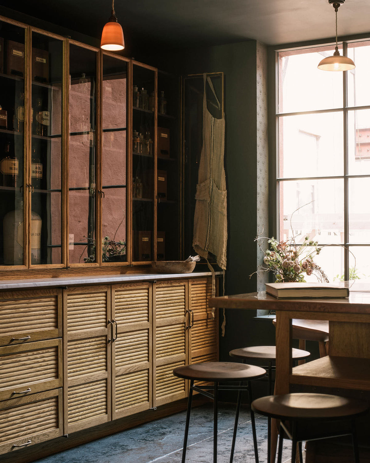 An oblique view of a cool and elegant Haberdasher’s collection features a large prep table with a copper worktop, a pantry run with tambour base cabinets, and glazed countertop cabinets. Adorned with vintage furniture and items, artisan ceramics, the deVOL porcelain light collection, Creedy stools, assorted containers, bottles, and a stunning bouquet of dried flowers, this space creates an atmospheric social bar.