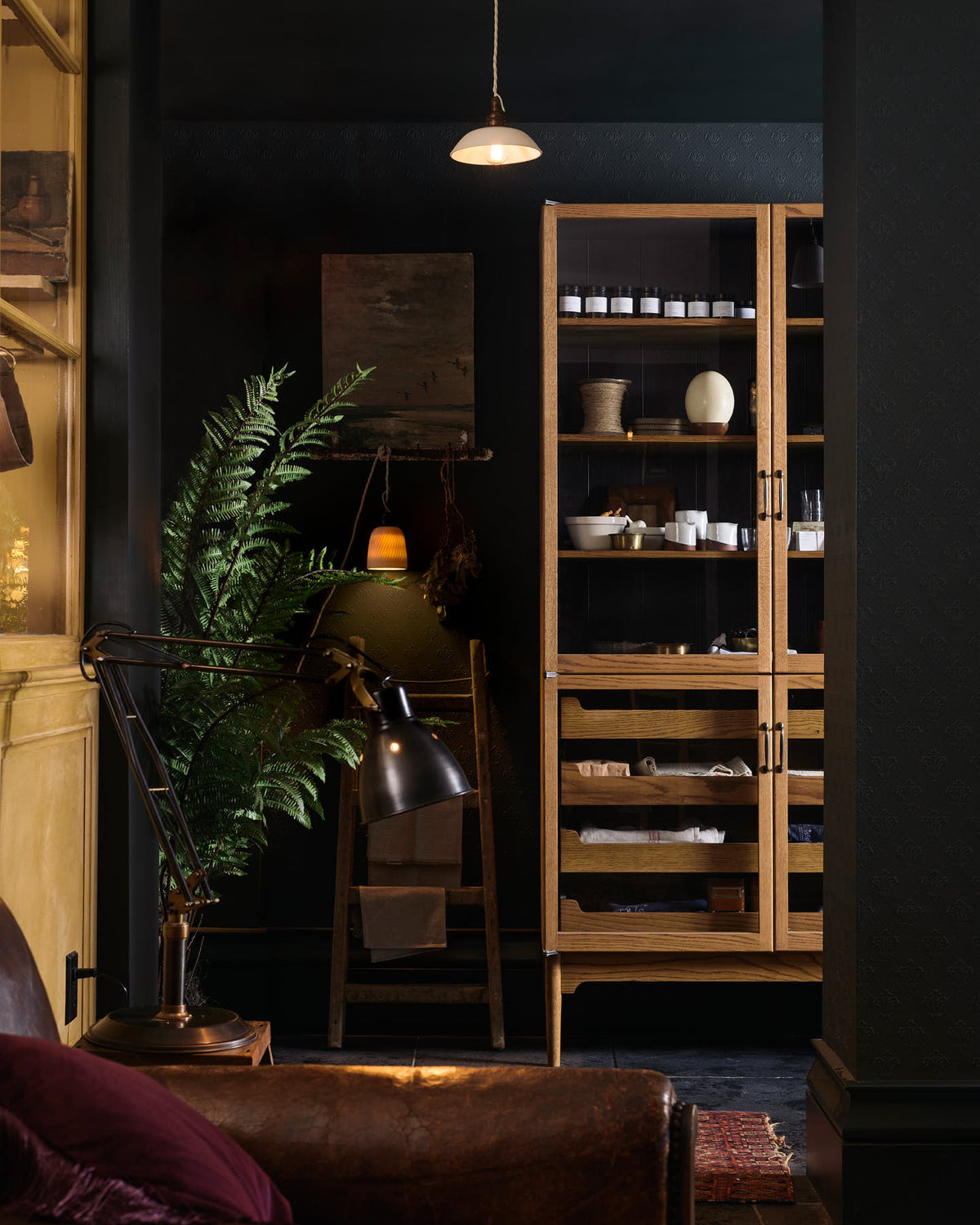 A quiet corner of a cool and elegant Haberdasher’s collection features a freestanding pantry with glazed doors. Adorned with vintage furniture and items, artisan ceramics, a plug-in porcelain beaker lights, assorted containers, and candles, this space creates an atmospheric social bar.