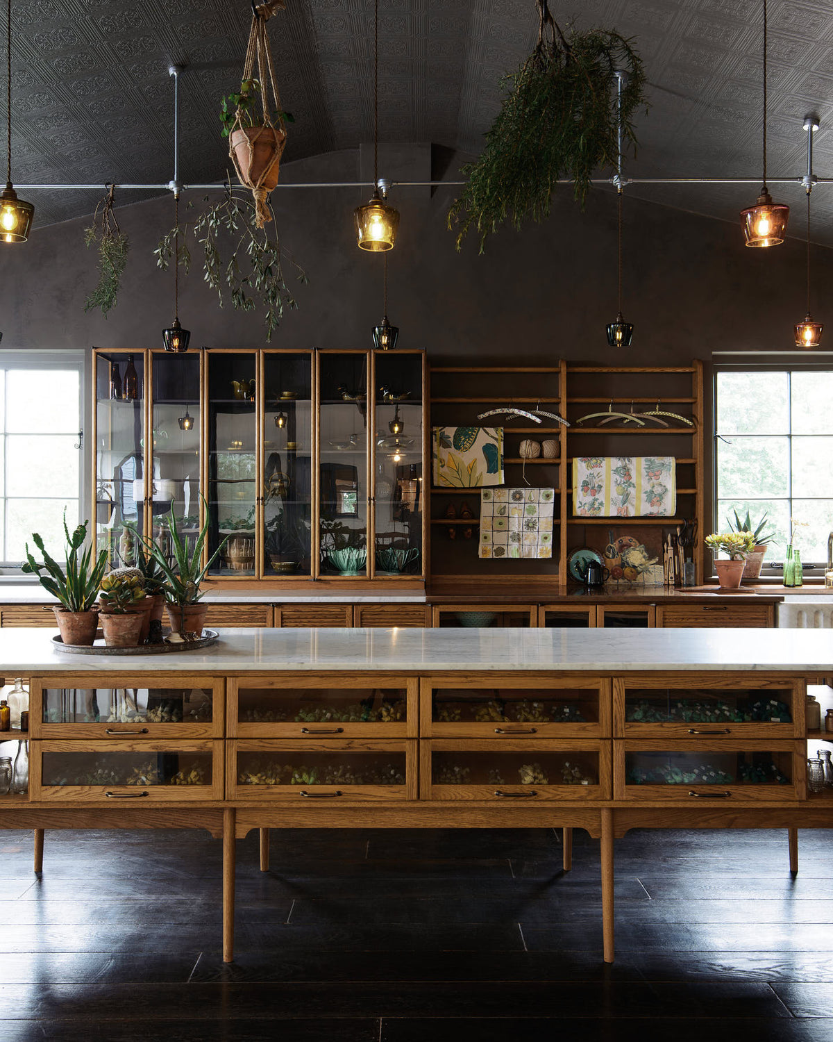 A warm and inviting Haberdasher’s kitchen features a long sink run with a Carrara marble and copper worktop, a double-fluted marble sink, a kitchen island, wall cupboards, and countertop cupboards. Adorned with luscious green hanging and potted plants, vintage items, artisan ceramics, illustrative tea towels, paintings, and a beloved white gas stove, this kitchen creates a lovely exhibitionist space that is both beautiful and practical.