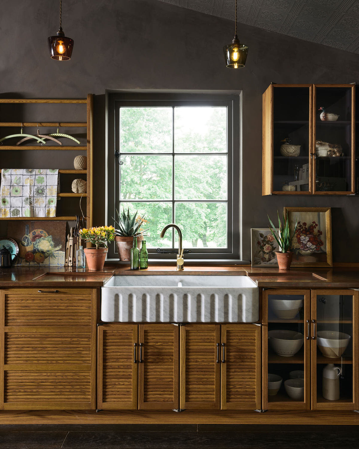 A closer look at a warm and inviting Haberdasher’s kitchen features a long sink run with a copper worktop, a double-fluted marble sink, a kitchen island, and wall cupboards. Adorned with luscious green potted plants, vintage items, artisan ceramics, illustrative tea towels, and paintings, this kitchen creates a lovely exhibitionist space that is both beautiful and practical.