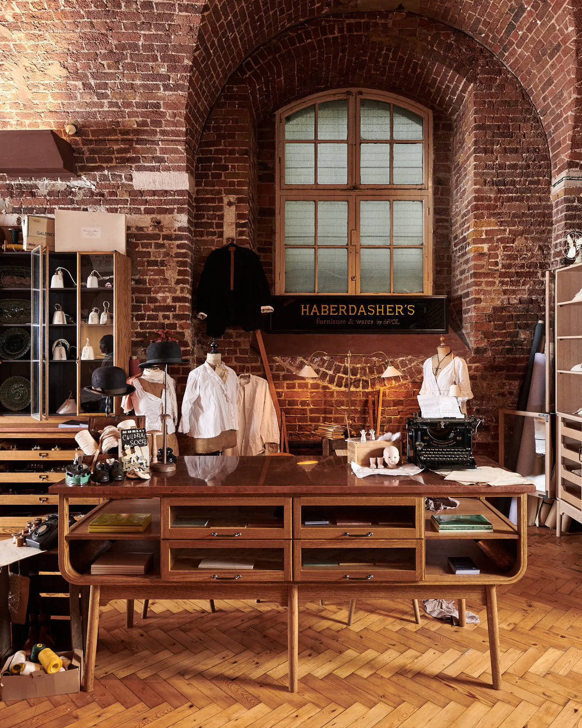 A handsome range of oak furniture and lighting from the deVOL Haberdasher’s collection features a kitchen island, countertop cupboards, and a glazed freestanding pantry. Adorned with deVOL Heirloom accessories, Helen stools, vintage furniture and items, dressmaker mannequins, and 1900s tailor tools, this space creates a beautiful homage to the English gentlemen’s haberdashery inspiration in a bricked wall showroom.