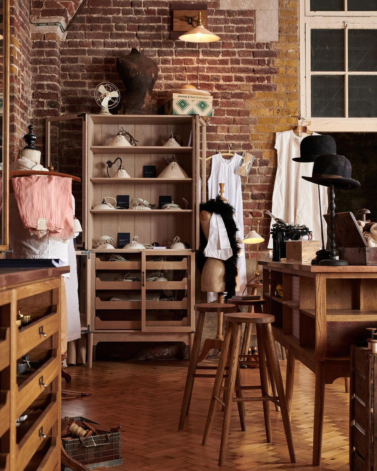 A handsome range of oak furniture and lighting from the deVOL Haberdasher’s collection features a kitchen island, countertop cupboards, and a glazed freestanding pantry. Adorned with deVOL Heirloom accessories, Helen stools, vintage furniture and items, dressmaker mannequins, and 1900s tailor tools, this space creates a beautiful homage to the English gentlemen’s haberdashery inspiration in a bricked wall showroom.