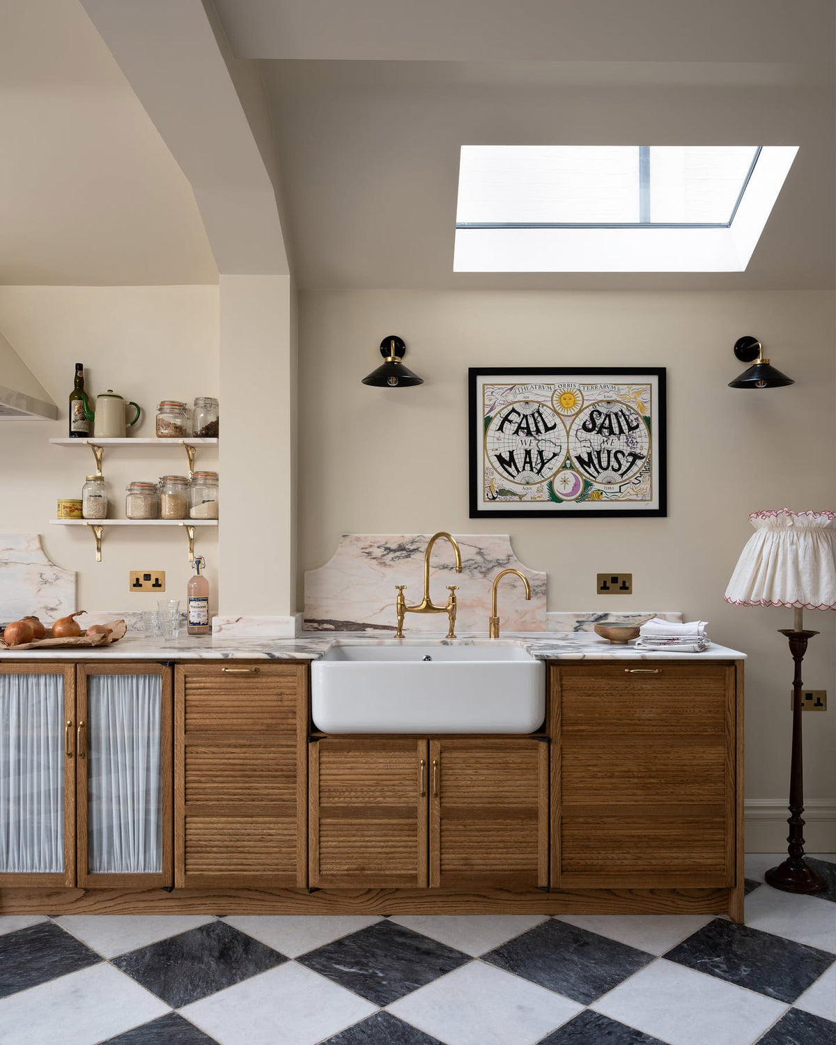 A grand and quaint Haberdasher’s kitchen features a sink and cooker run with a  marble worktop and decorative vanity cut, a single Belfast sink, and an aged brass Ionian tap. Beautifully decorated with a white gas cooker with golden accents, a matching canopy, a deVOL grand tour desk lamp, vintage furniture, and a cute black cat lying on the chequerboard flooring, this kitchen creates a humble and functional space with a touch of elegance.