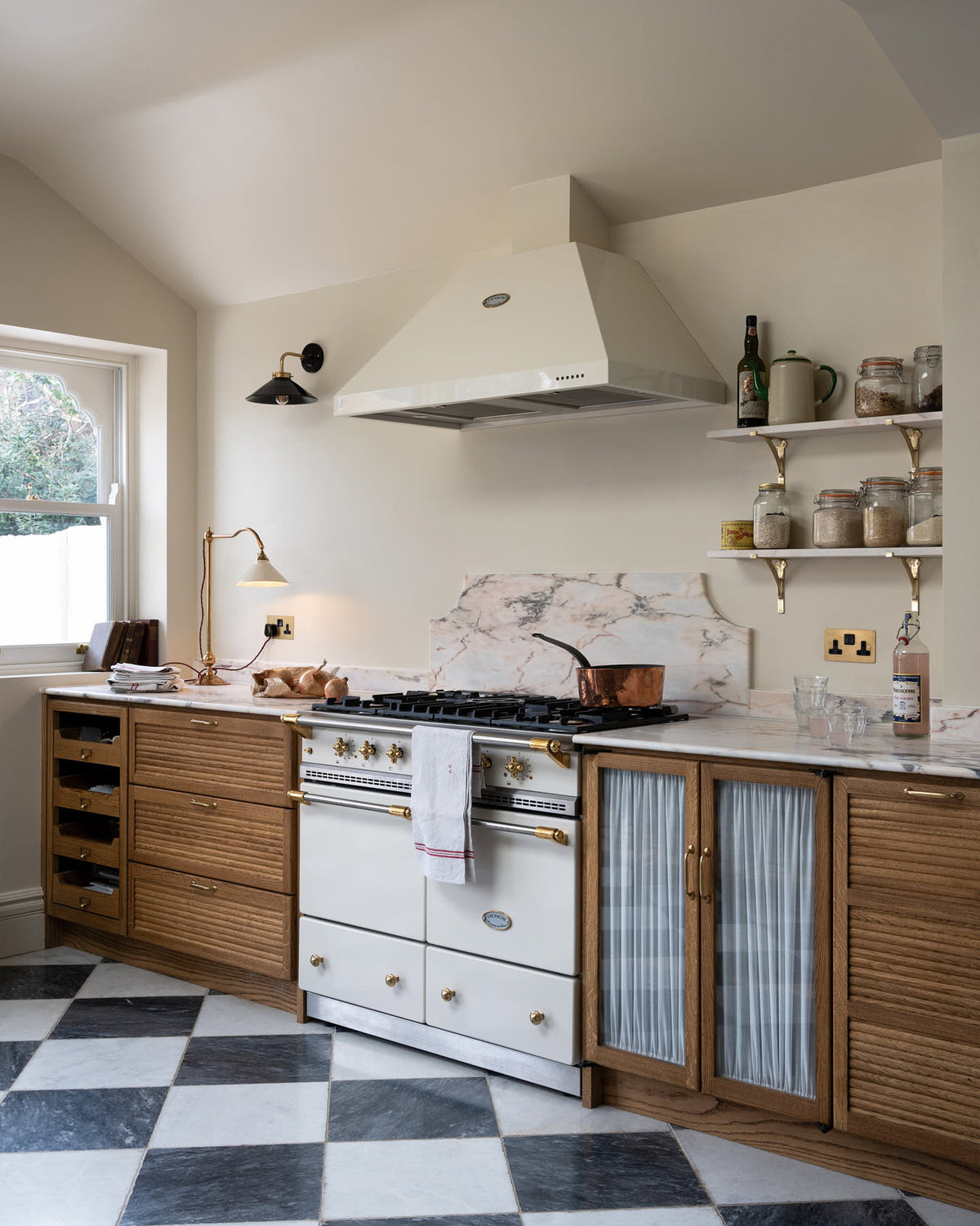 An oblique view of a grand and quaint Haberdasher’s kitchen features a sink and cooker run with a Paonazzo marble worktop and decorative vanity cut. Beautifully decorated with a white gas cooker with golden accents, a matching canopy, a deVOL grand tour desk lamp, vintage furniture, beloved chapel chairs, a skylight, and a cloth lampshade, this kitchen creates a humble and functional space with a touch of elegance.