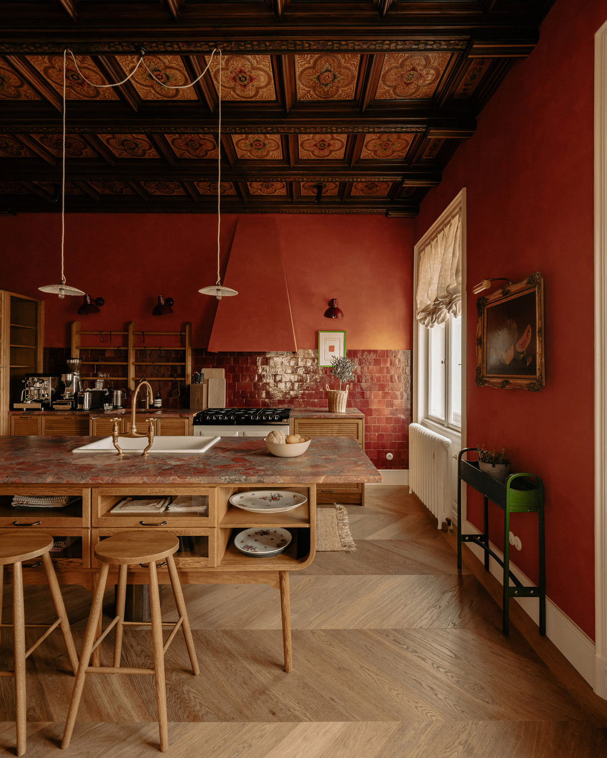 A warm and historical Haberdasher’s kitchen features a sink and cooker run with red Zellige splashback tiles, a kitchen island with an aged brass Ionian tap, and a red marble worktop. Beautifully decorated with decorative floral ceiling tiles, large narrow pleat porcelain lights, Helen stools, artisan ceramics, vintage items, and paintings, this kitchen creates a grand yet comfortable space.