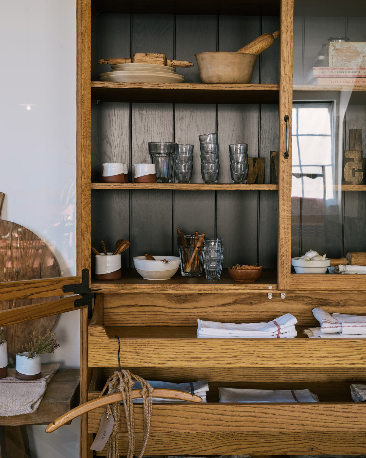 A closer look at a warm and spacious Haberdasher’s kitchen featuring a freestanding pantry with glazed doors. Adorned with vintage furniture and items, artisan ceramics, drinking glasses, tea towels, large wooden tapas boards, and wooden letterpress, this kitchen creates a contemporary yet nostalgic space.