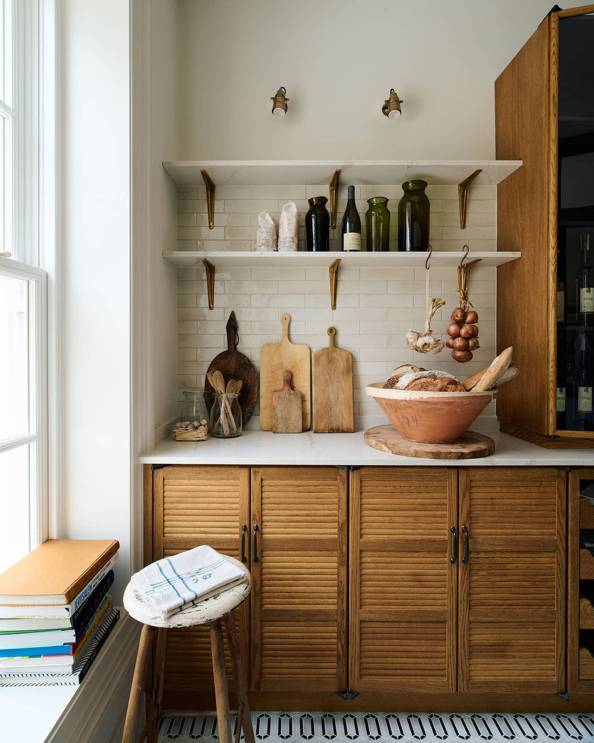 A smart and functional Haberdasher’s pantry features a long pantry run with glazed, open cupboards with internal drawers and tambour doors, a Carrara marble worktop and shelves, and cream splash back tiles. Beautifully decorated with eye-catching diamond mosaic flooring, oxidised brass hooks, cloves of garlic, onions, loaves of bread, and classic shelf brackets, this pantry creates a warm and neat storage space.