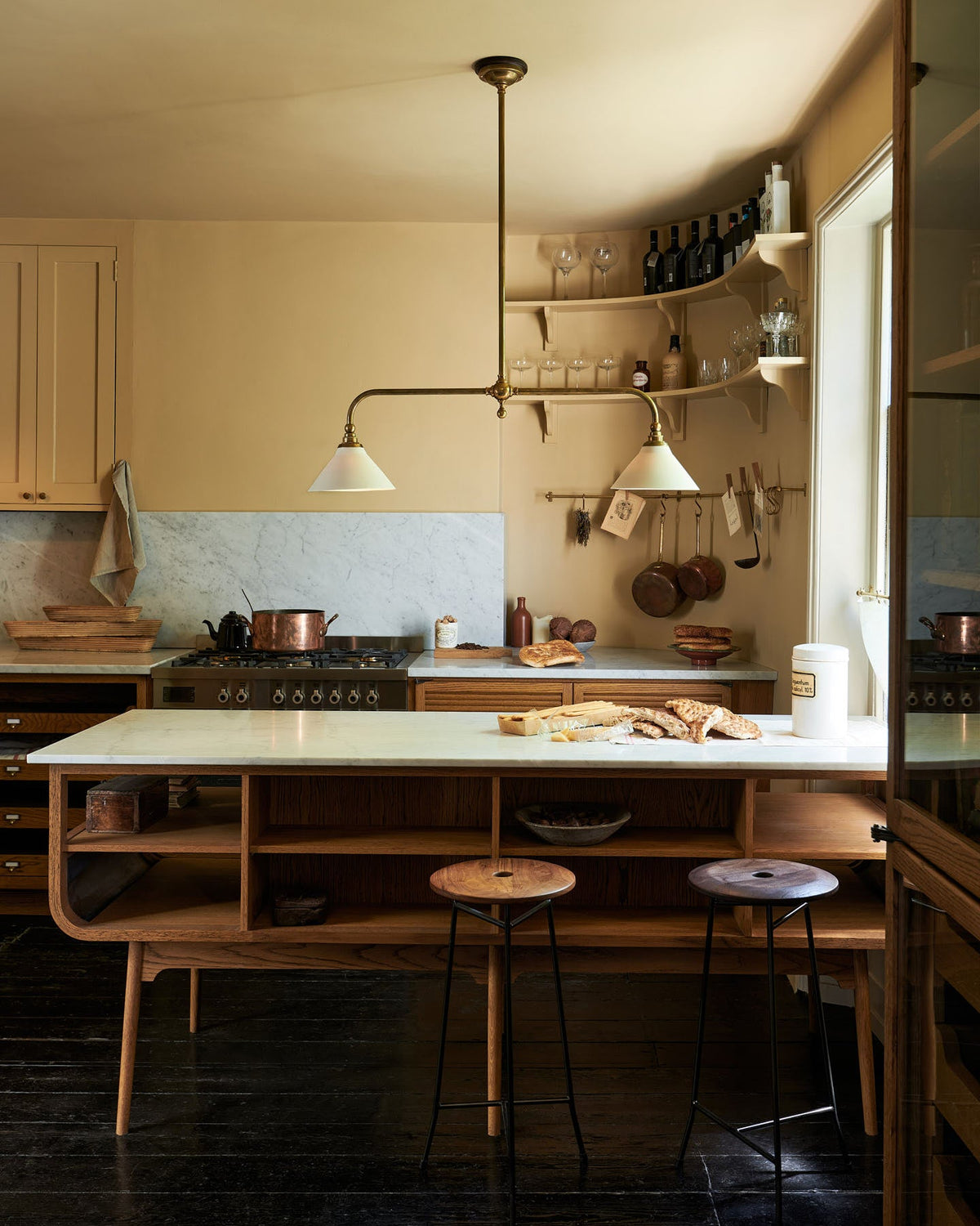 A cosy and sophisticated Haberdasher’s kitchen features a cooker run with a Carrara marble worktop, a small prep table, bespoke curved shelves, and muted yellow walls. Beautifully decorated with Creedy stools, a deVOL Heirloom gaselier light, Heirloom wall lights, aged brass hanging rails, copper pans, focaccia bread, vintage items, and artisan ceramics, this kitchen creates a bright and warm space.