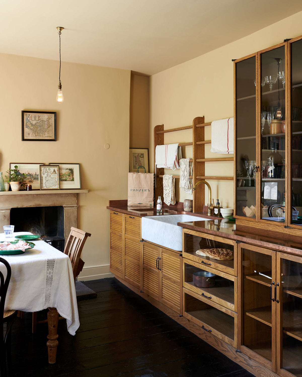 An oblique view of a cosy and sophisticated Haberdasher’s kitchen features a sink run with a copper worktop, a single marble sink, glazed countertop cupboards, and muted yellow walls. Beautifully decorated with Bentwood café chairs, vintage furniture and items, framed detailed illustrations, lace runners, tea towels, and artisan ceramics, this kitchen creates a bright and warm space.