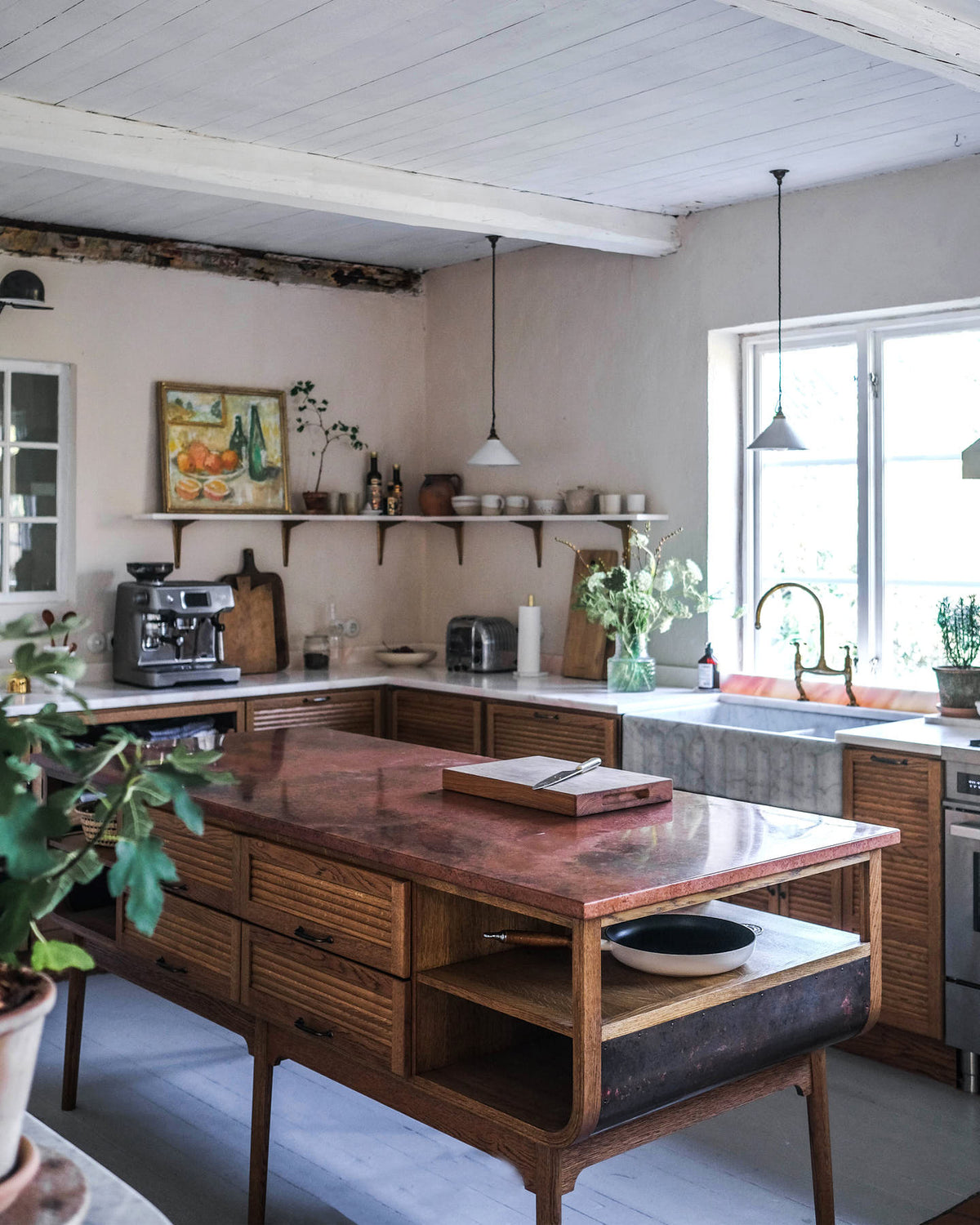 An oblique view of a warm and inviting Haberdasher’s kitchen features a sink and cooker run with a Carrara marble worktop, a double-fluted marble sink, an aged brass Ionian tap, prep tables with a copper worktop, and a shelf. Beautifully decorated with vintage furniture and items, artisan ceramics, luscious green potted plants, and a still life painting, this kitchen creates an airy and cosy Scandinavian small fishing town hideaway.