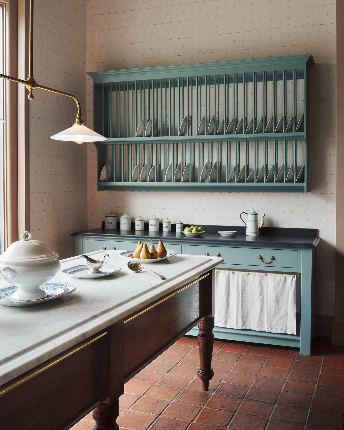 An oblique view through a doorway of a beautiful and functional Trinity blue Heirloom kitchen features open cabinets, plate racks, and a showpiece Dairy table. Lovingly adorned with bone-white artisan ceramics, selected antiques, lace curtains, Gaselier lights, and earthy red terracotta square floor tiles that recreate a Victorian kitchen made to last a lifetime.