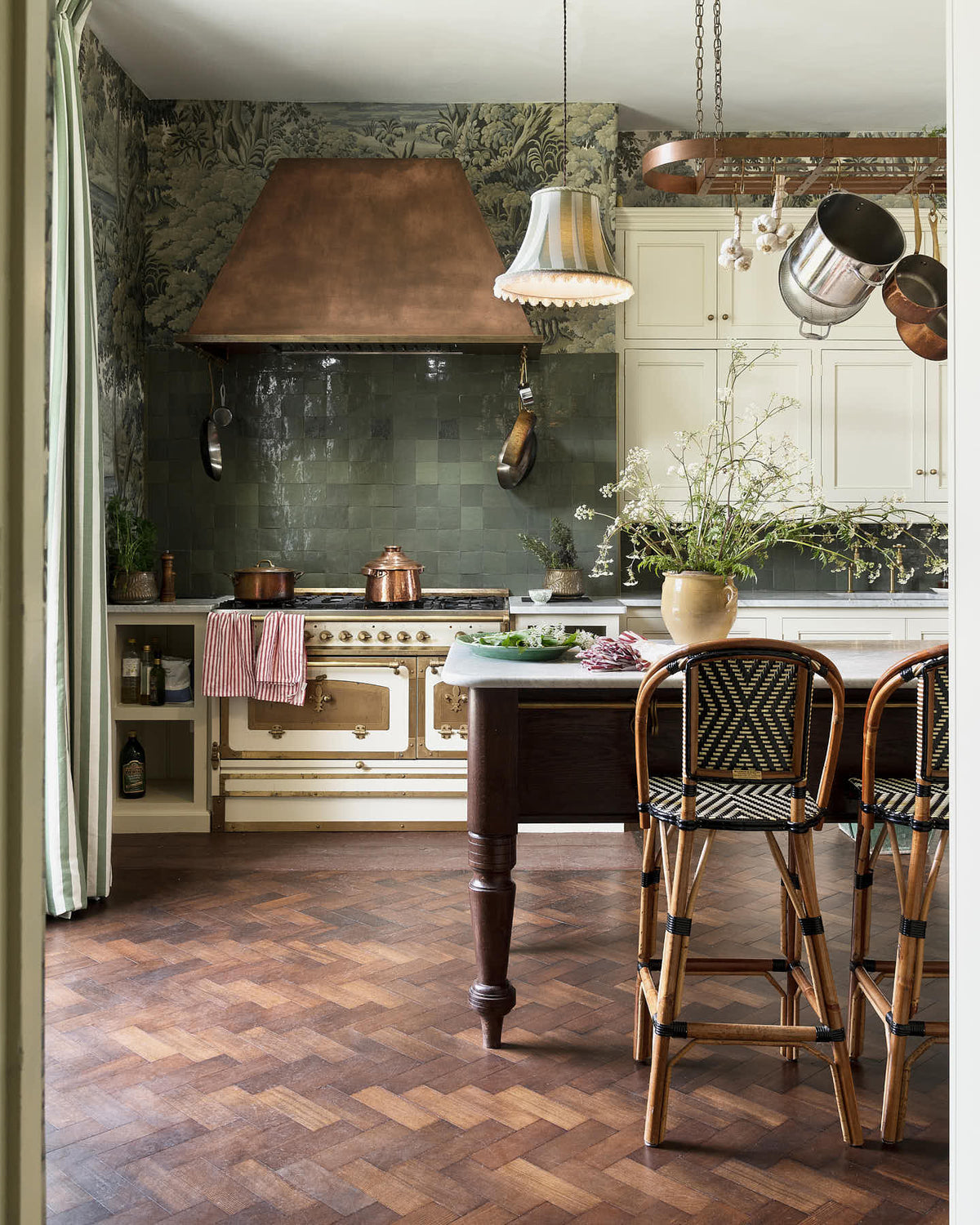 A grand and eclectic kitchen features classic English cupboards with castellations, a Carrara marble worktop, and a stunning Heirloom dairy table. Beautifully decorated with detailed muted-green rainforest wallpaper, handmade Zellige tiles, a white and gold Italian gas cooker, a copper hood, and a white wildflower floral arrangement, this kitchen creates a fantasy experience in a castle.