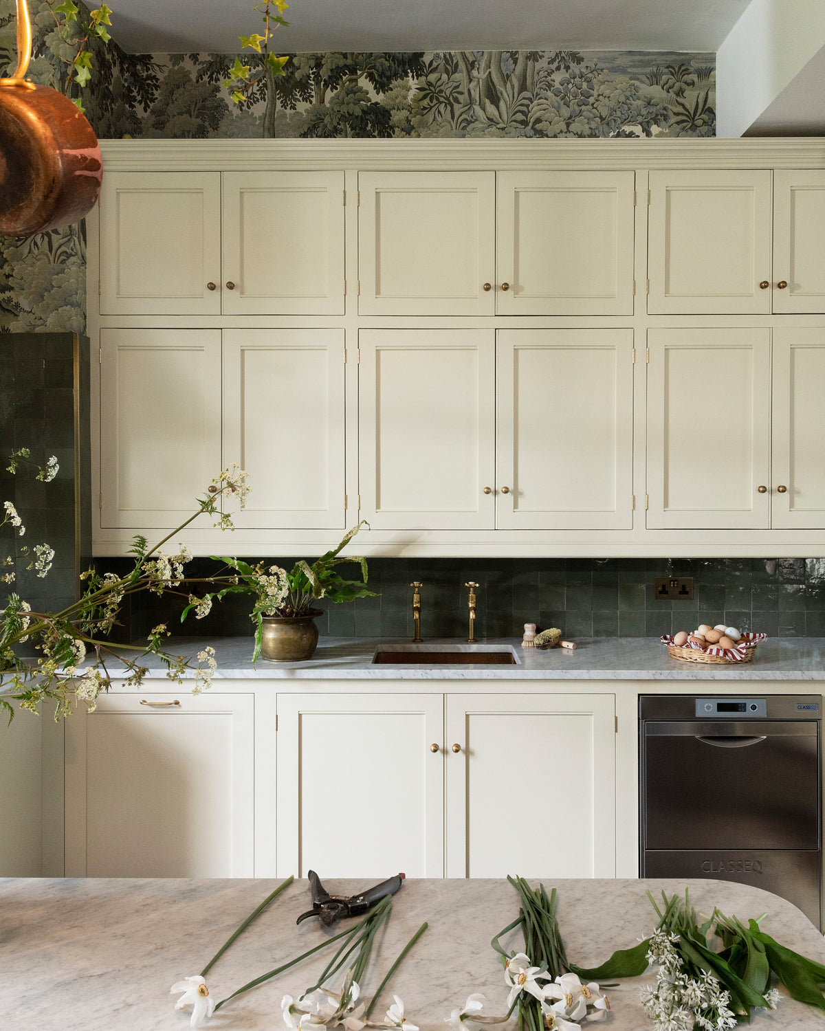 A grand and eclectic kitchen features a classic English sink run with a single copper sink, aged brass Mayan taps, wall cupboards, a Carrara marble worktop, and a stunning Heirloom dairy table. Beautifully decorated with detailed muted-green rainforest wallpaper, copper pans, a basket of fresh eggs, and a white wildflower floral arrangement, this kitchen creates a fantasy experience in a castle.