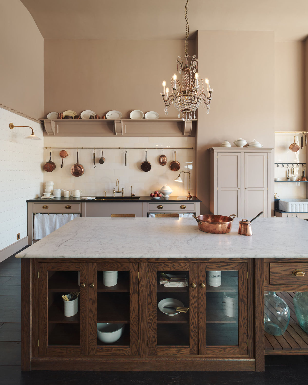 A grand Princelet pink Heirloom and Classic English kitchen features a sink cabinet with a black granite worktop, a butler’s pantry, a large kitchen island with a Carrara marble worktop, aged brass rails, ditsy delft wallpaper, a tall cream subway tile splashback, a grand tour desk lamp, and an Heirloom task light. Lovingly adorned with brass pots and pans, artisan ceramics, and an antique crystal chandelier, creating a warm, delicate, and airy kitchen space.